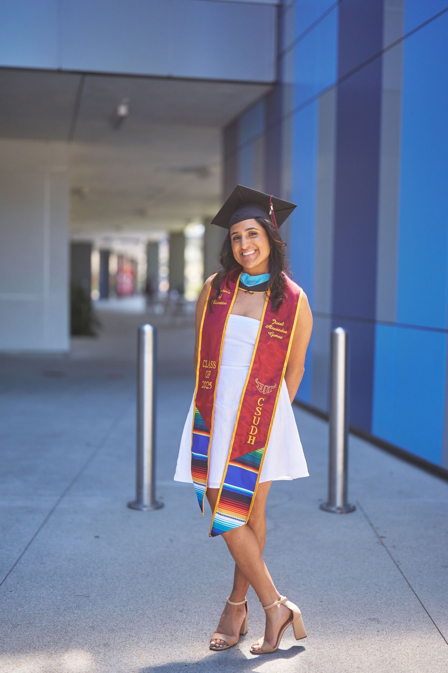 Graduate in white dress and red stoles poses confidently in cap and gown.