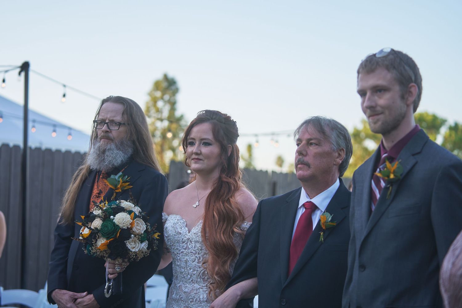 Outdoor wedding ceremony with bride in white gown holding bouquet, flanked by three men in formal attire.