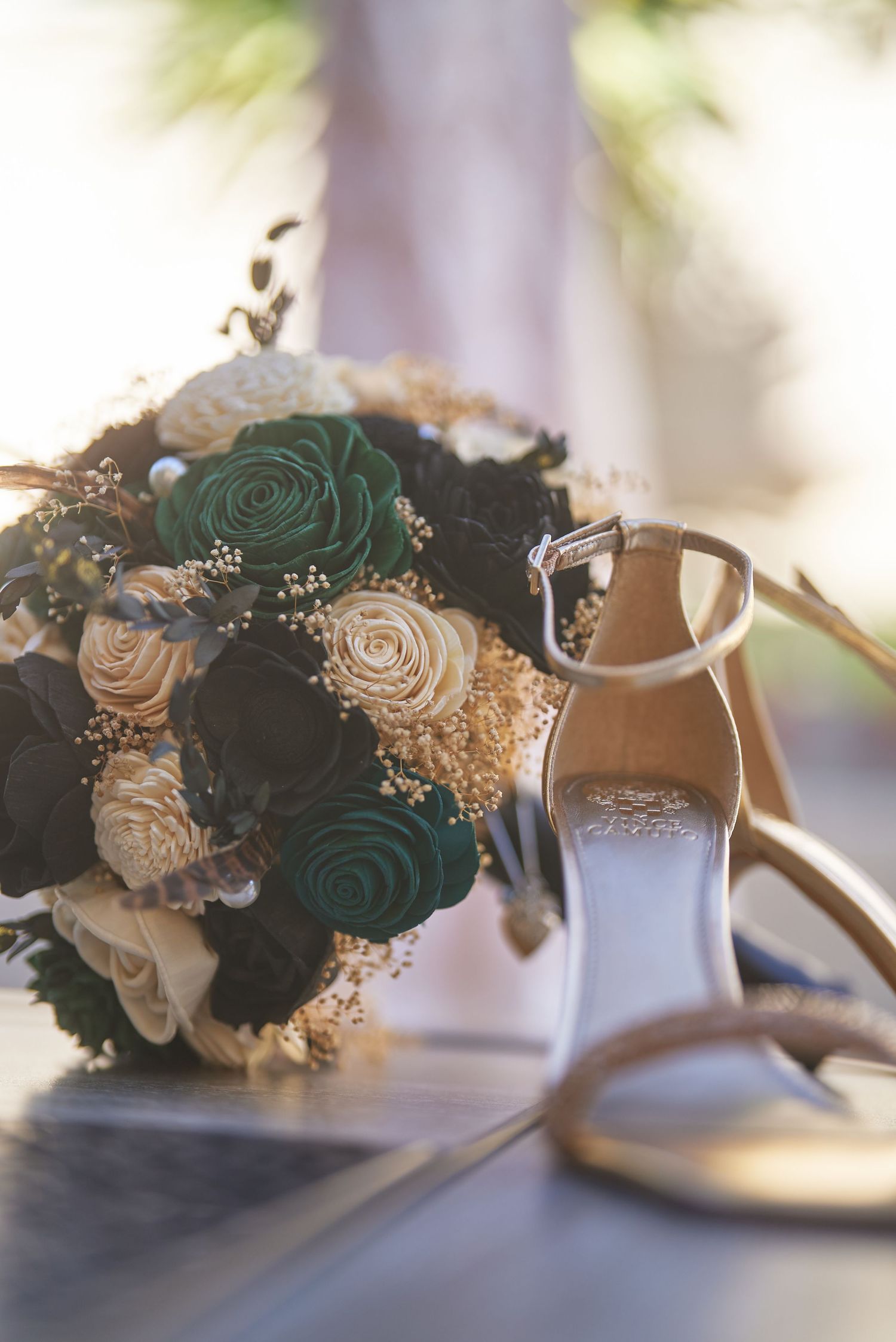 Elegant bridal bouquet with green and ivory roses beside gold ankle-strap heels on a marble surface.