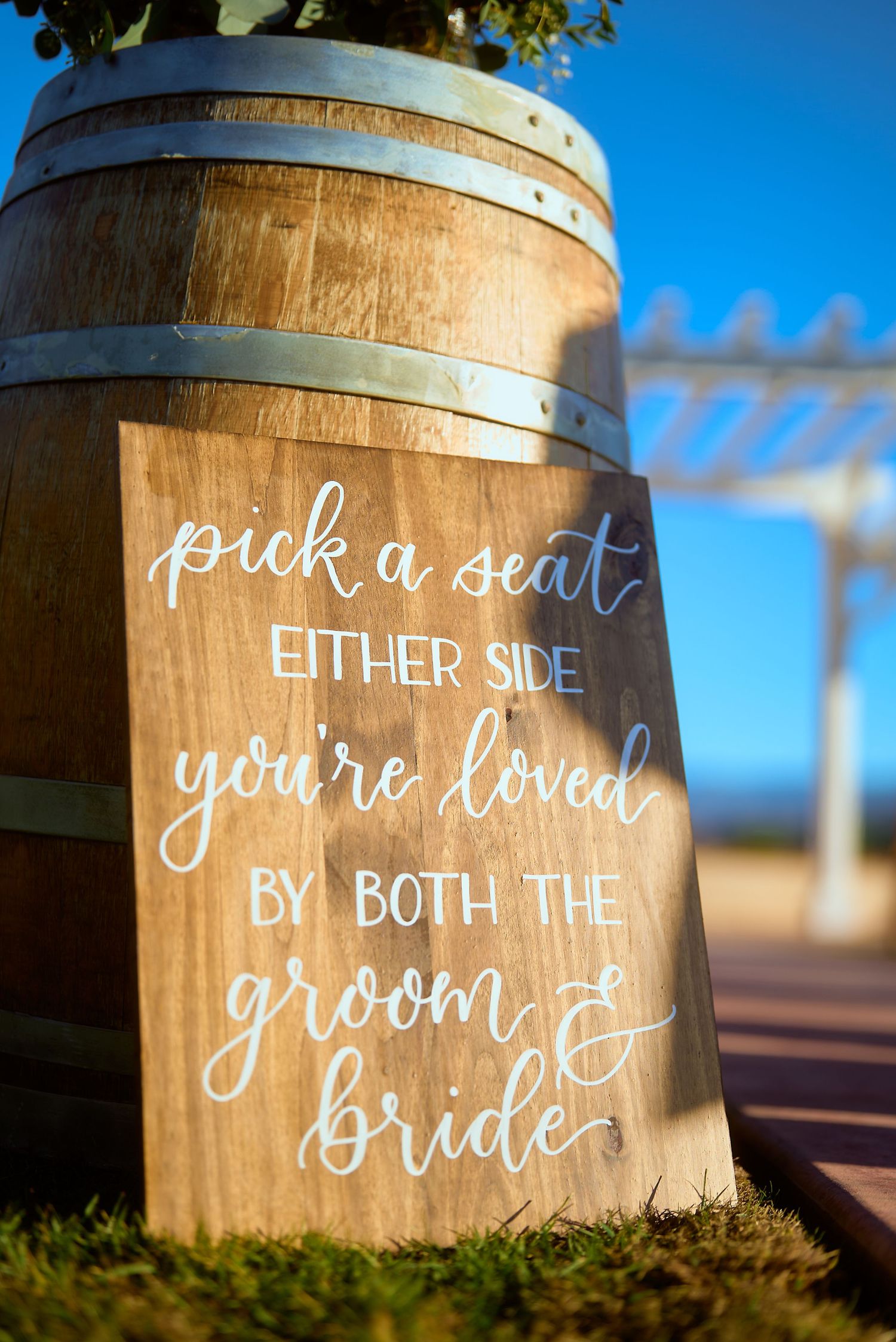 Rustic wooden wedding sign leaning on wine barrel reads pick a seat either side you're loved by both the groom and bride.