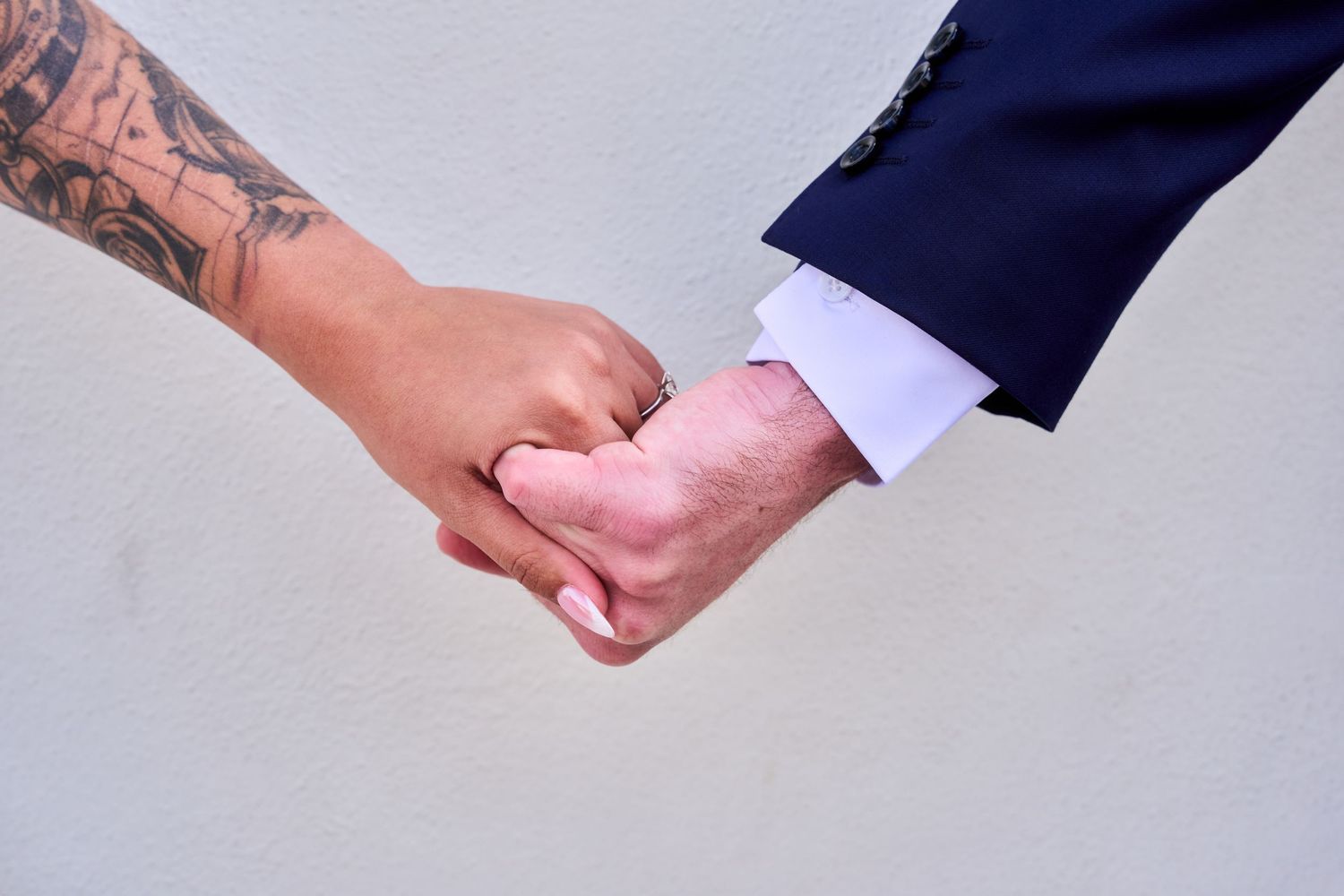 Close-up of two people holding hands, one with a tattoo sleeve, the other in a navy suit, symbolizing a wedding couple.