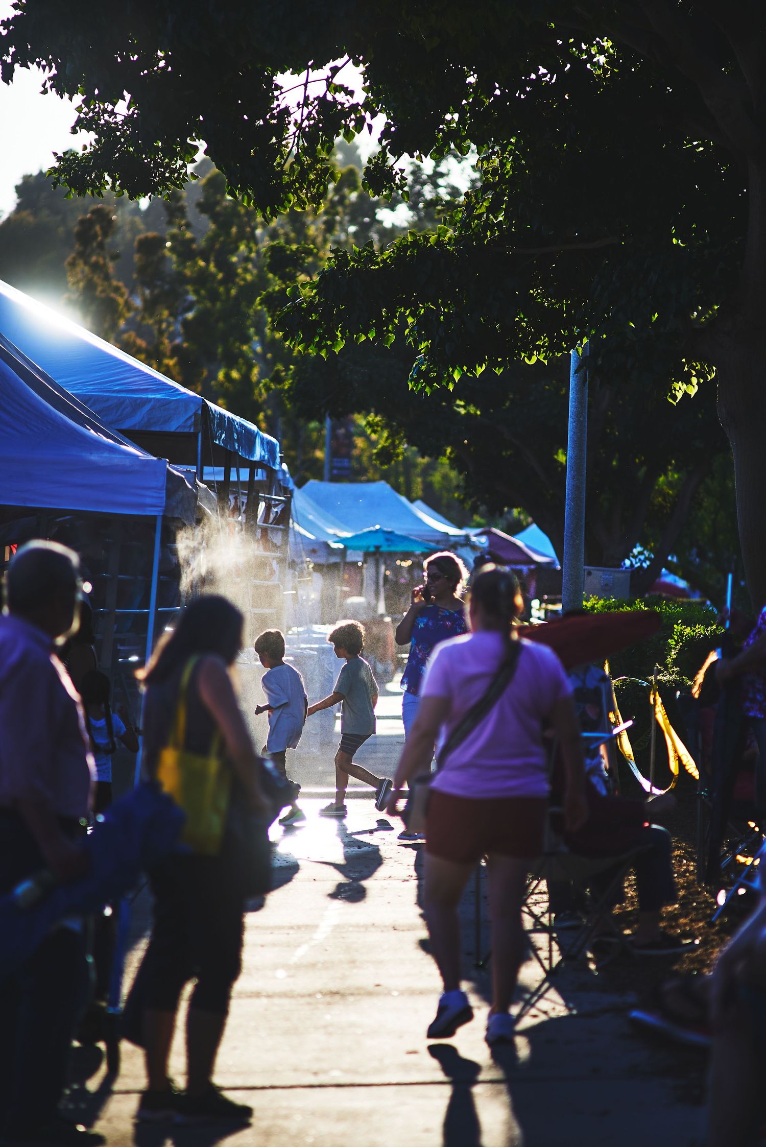 Silhouettes of people walking through outdoor market with blue tents and sunlight streaming.