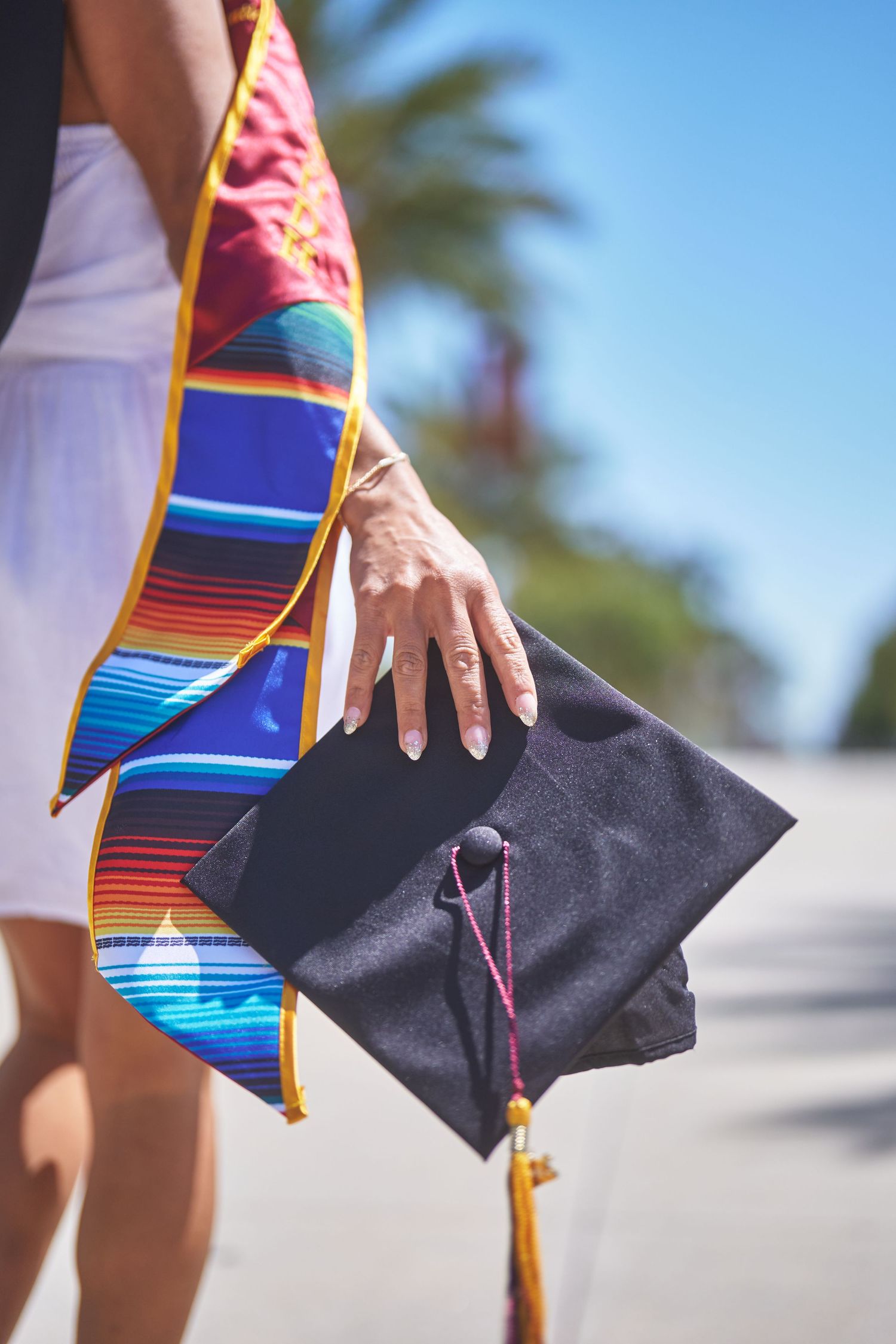 Graduate holding black mortarboard with colorful serape graduation stole on sunny campus day.