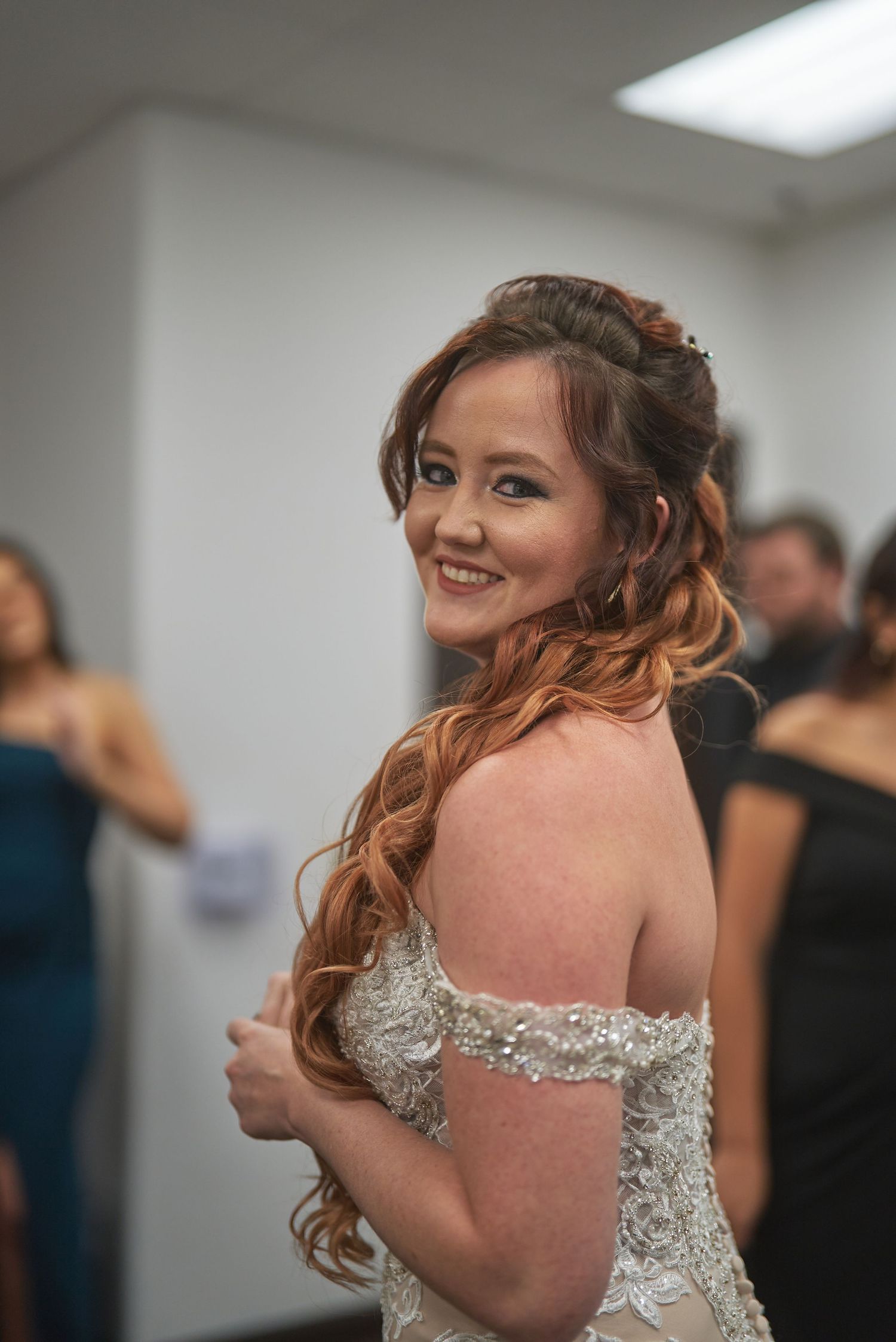 Smiling bride in beaded off-shoulder wedding gown with auburn curls, looking back over shoulder at wedding venue.