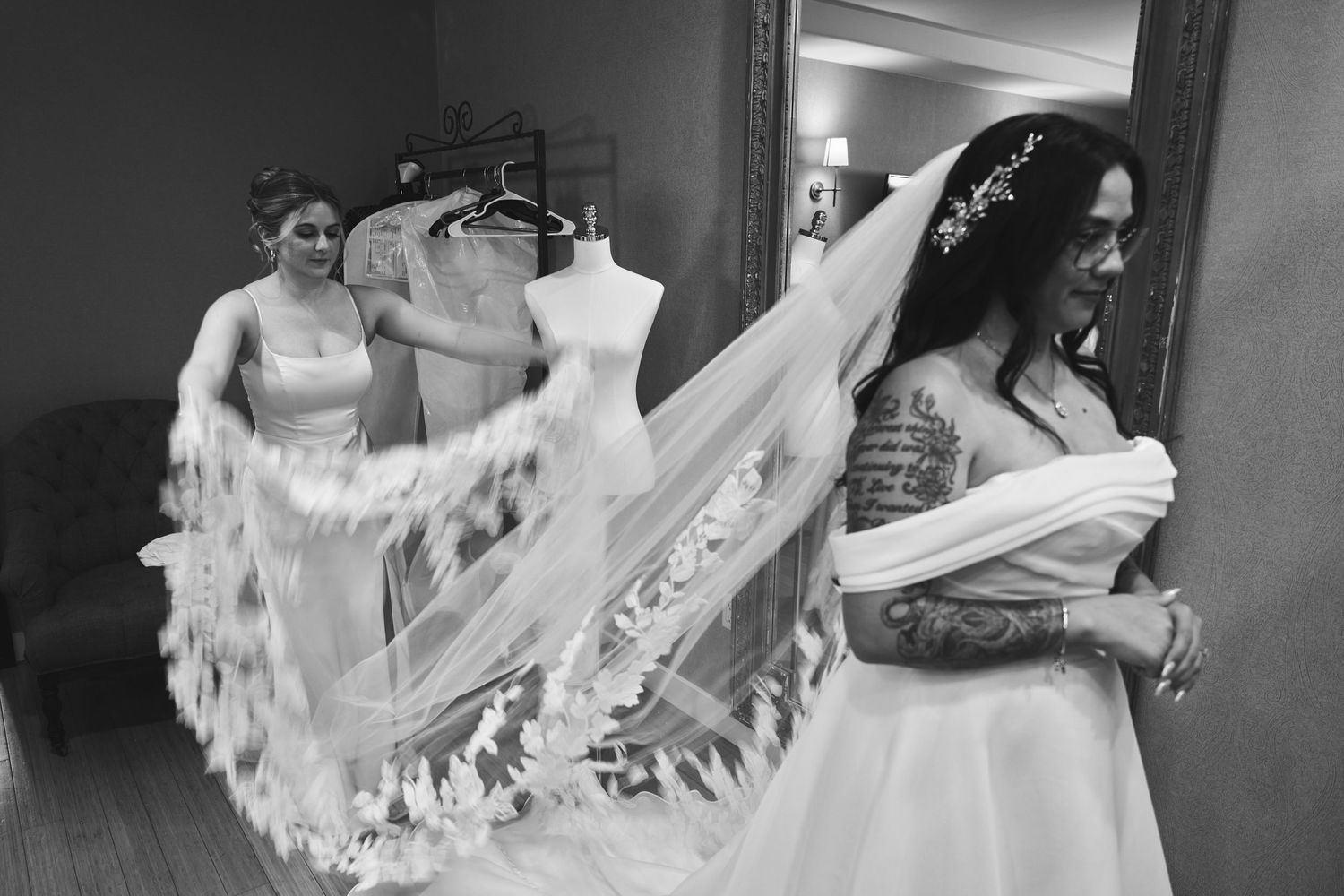 Black and white photo of tattooed bride holding veil as bridesmaid helps arrange it in a wedding preparation room.