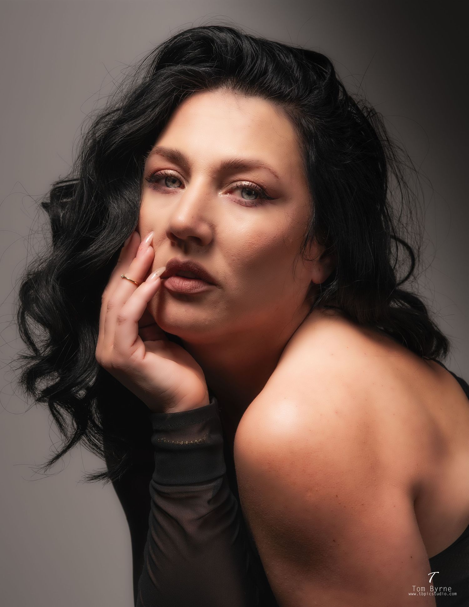 A dark-haired woman poses thoughtfully with her hand near her face against a gray studio background.