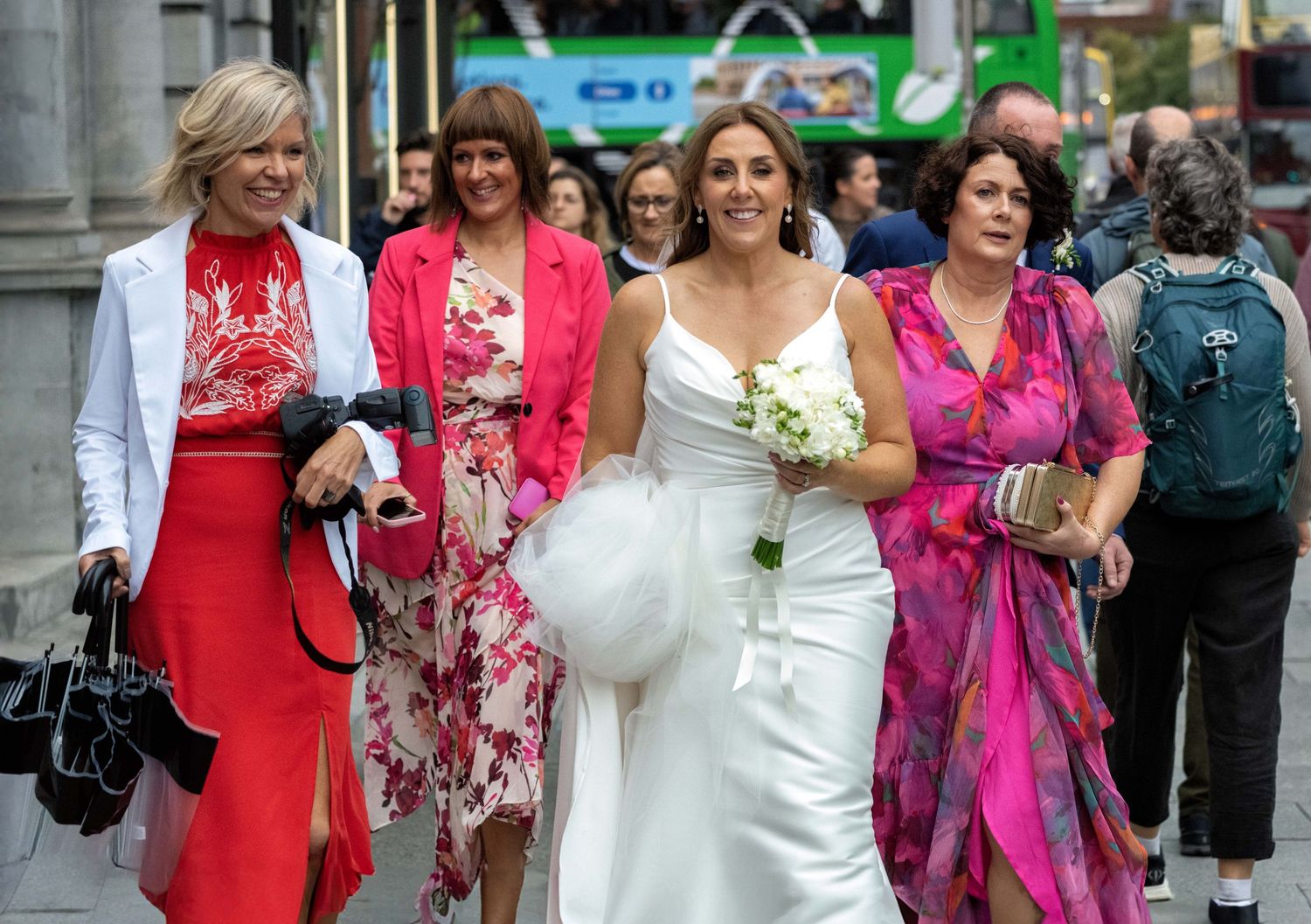 Bride in white gown holding bouquet walks with bridesmaids in colorful dresses on a busy city street.