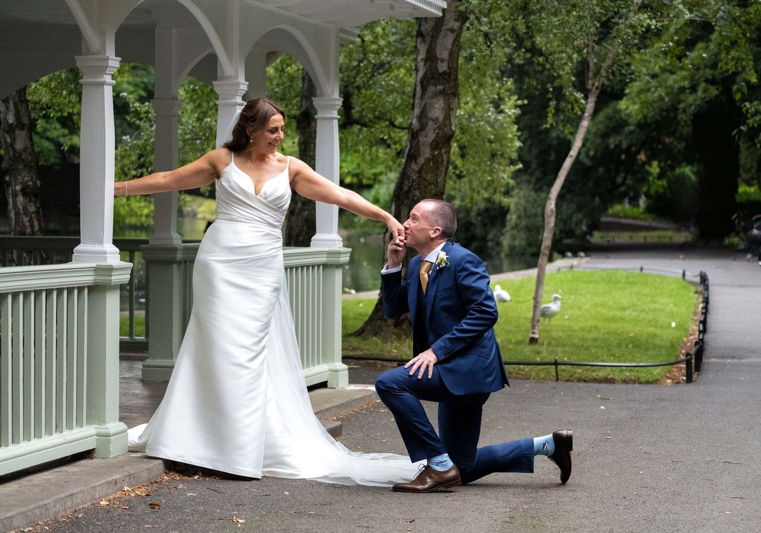 Groom in blue suit kneels kissing bride's hand in white gown at a gazebo during outdoor wedding ceremony.