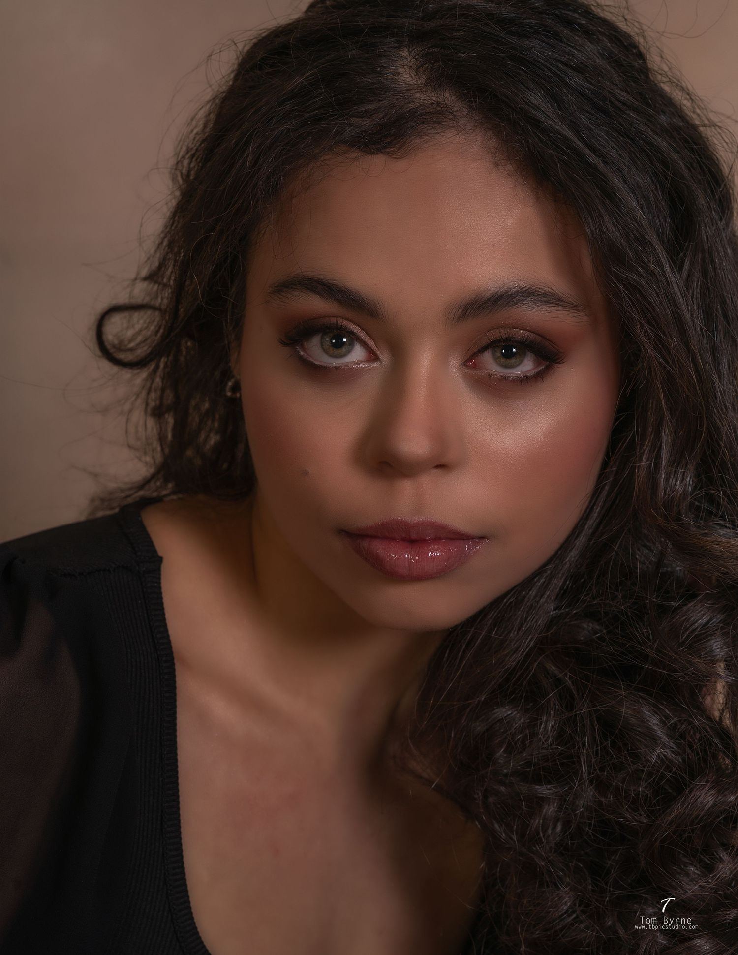 Close-up portrait of a young woman with curly dark hair, green eyes, and elegant makeup in professional studio lighting.