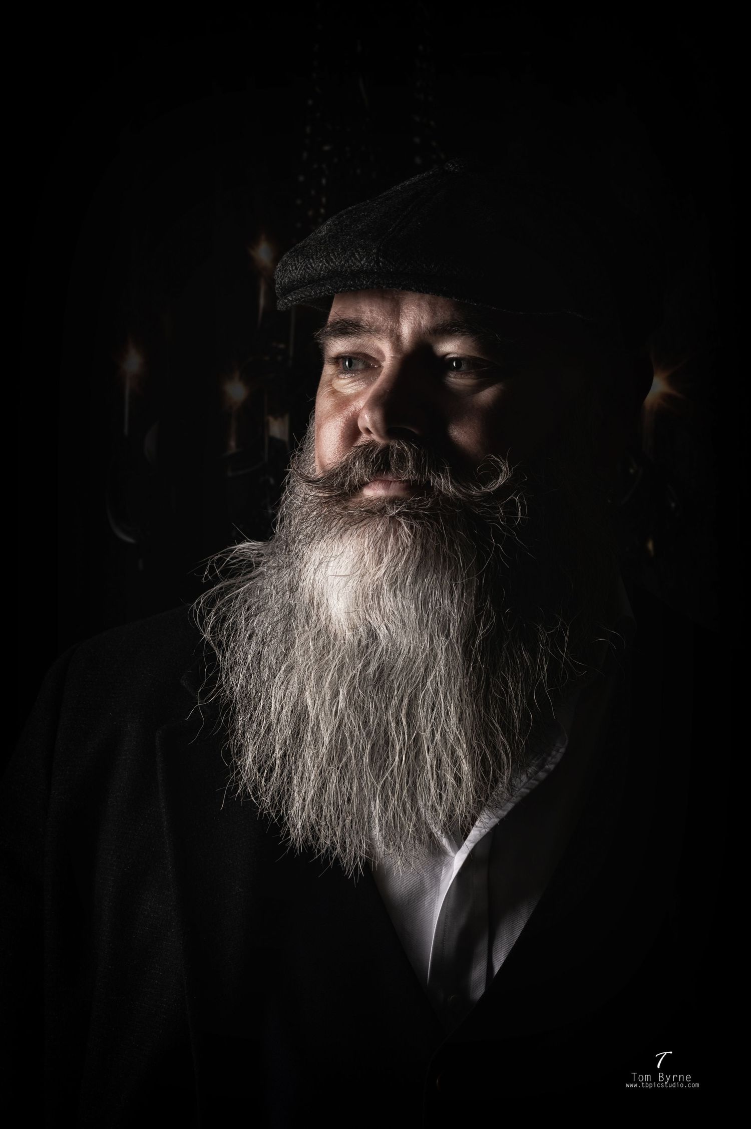 Dramatic portrait of an elderly bearded man with gray beard wearing dark hat, lit against black background.