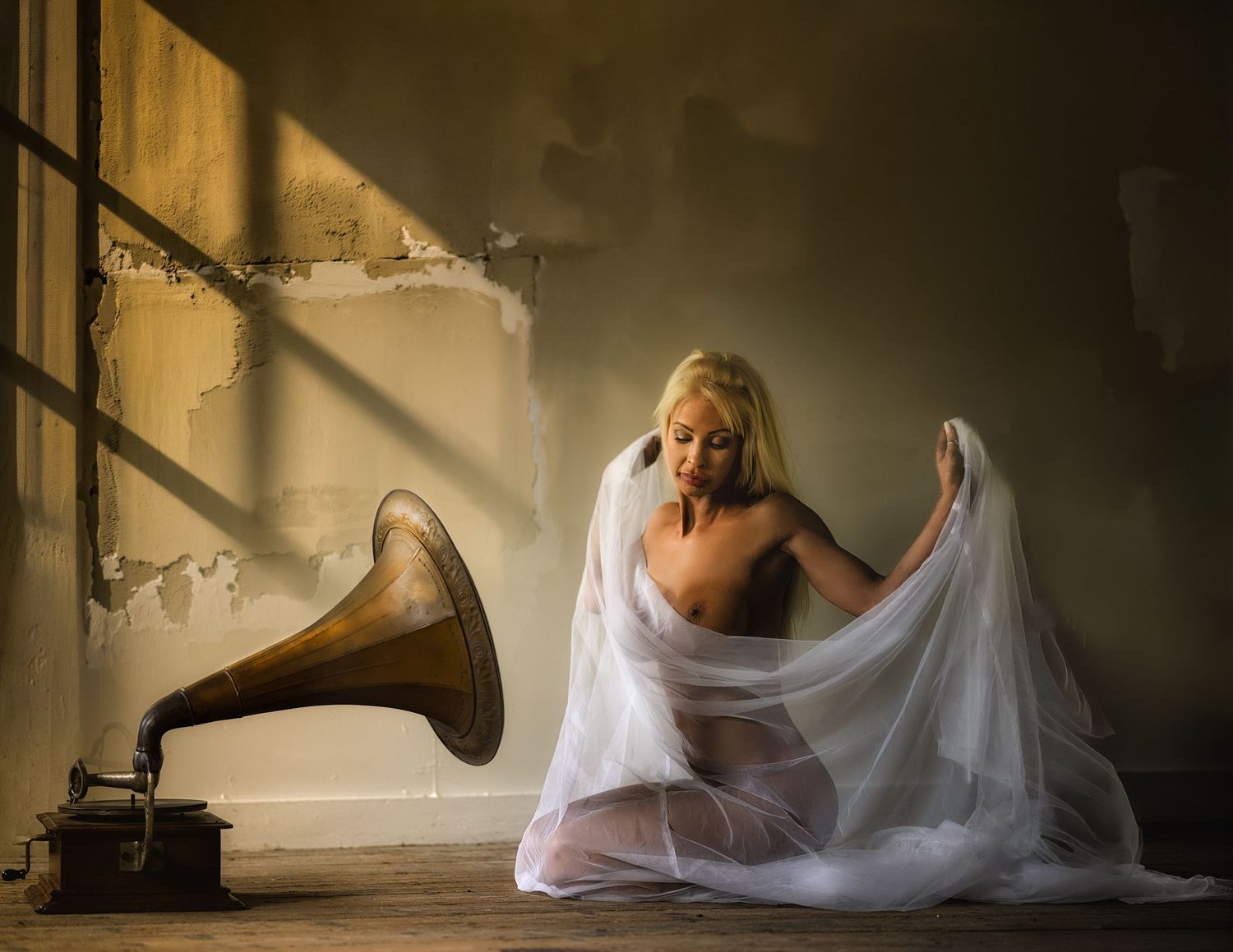Blonde woman draped in sheer white fabric kneels beside a vintage gramophone in a rustic, sunlit room.