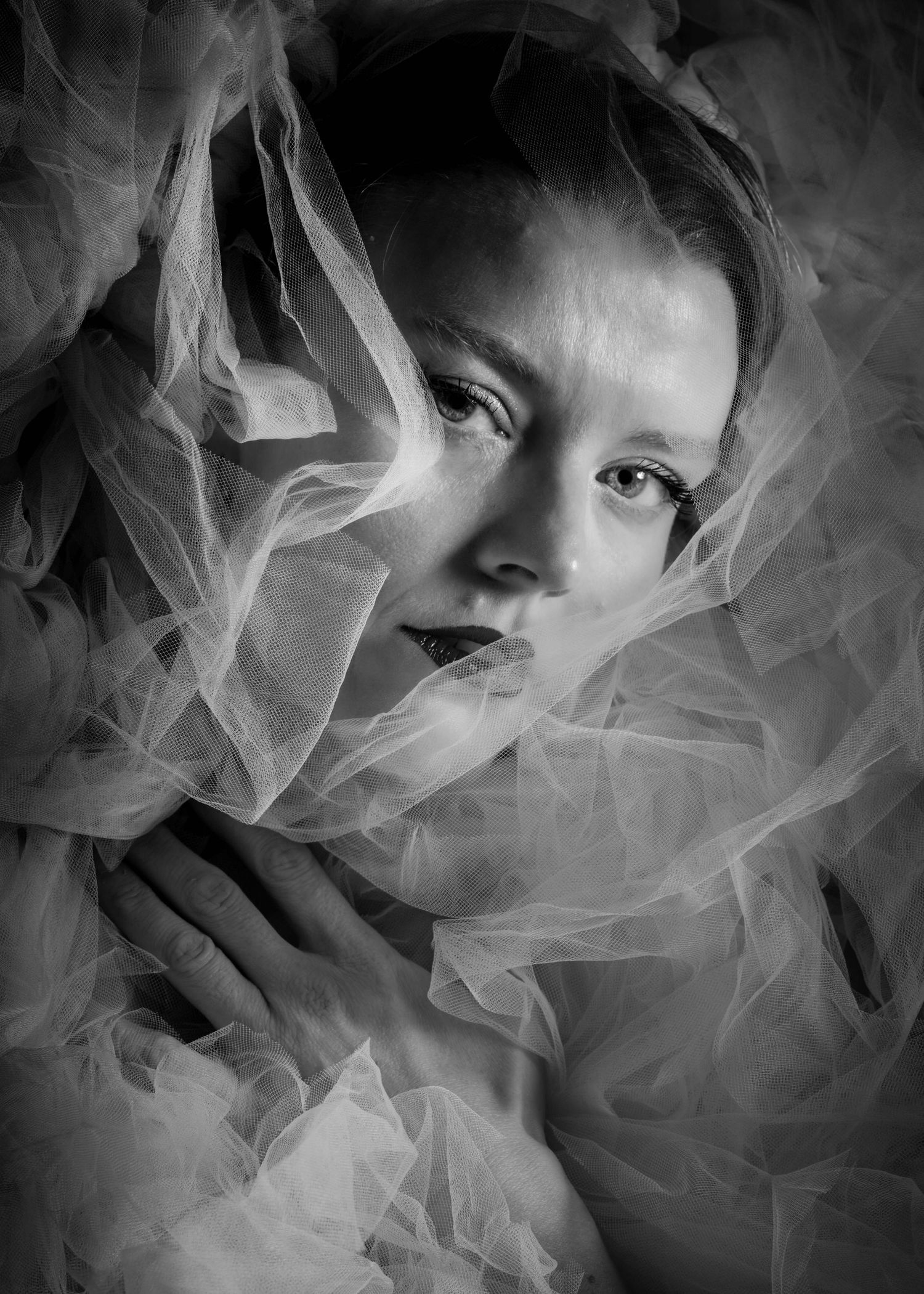 Black and white portrait of a young woman wrapped in sheer tulle fabric, gazing softly at the camera.
