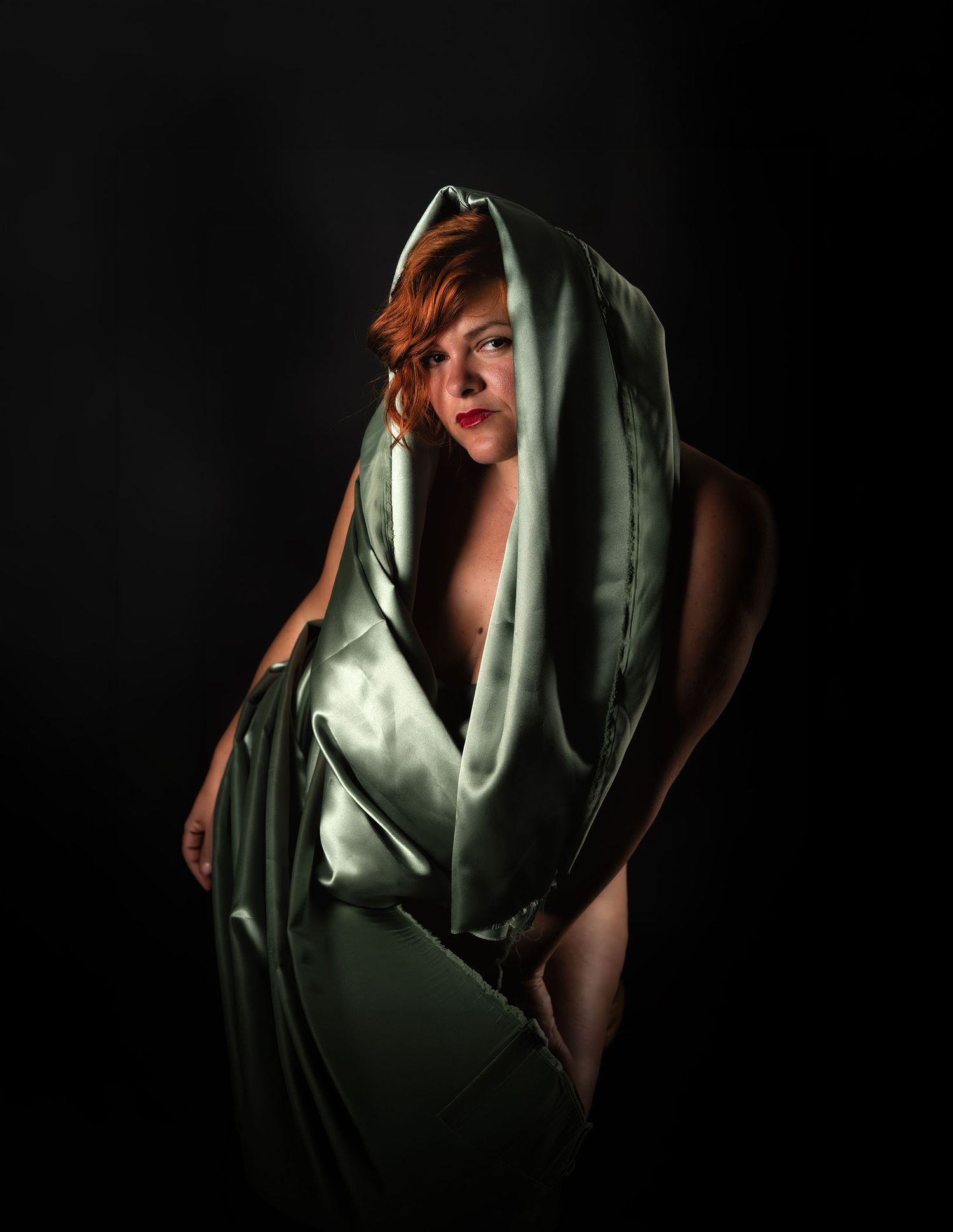 A red-haired woman draped in flowing green satin fabric against a dark background.