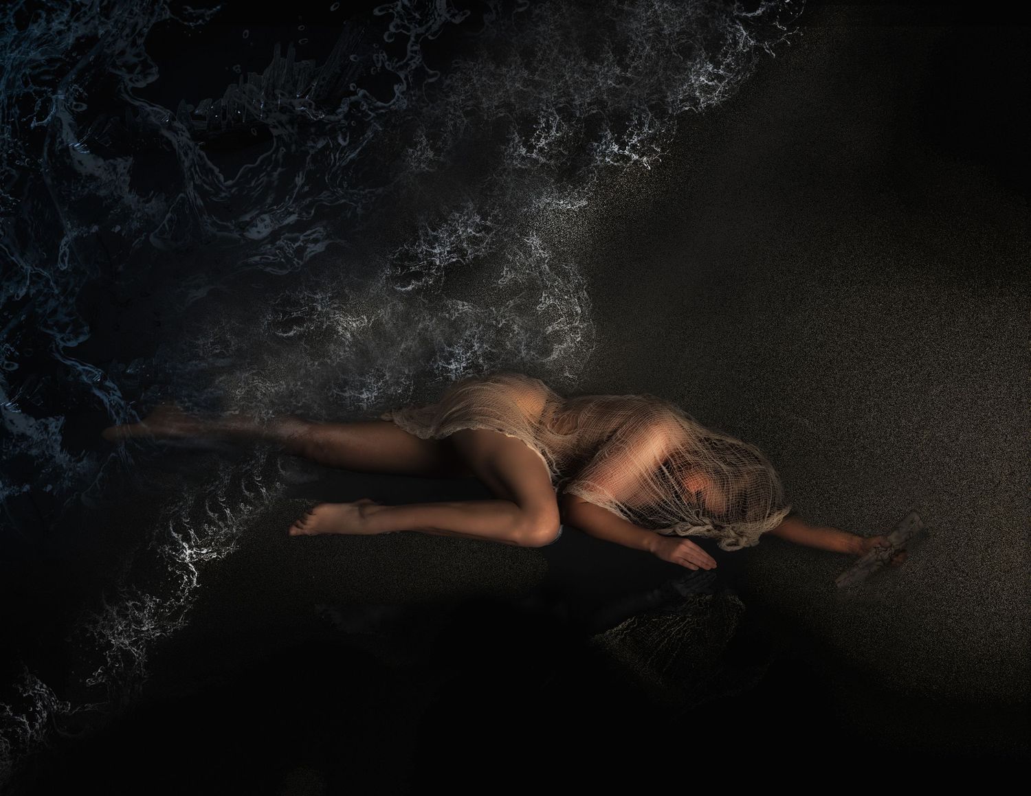 A woman in a bikini lies on a dark sandy beach as ocean waves wash over her in a dramatic aerial view.