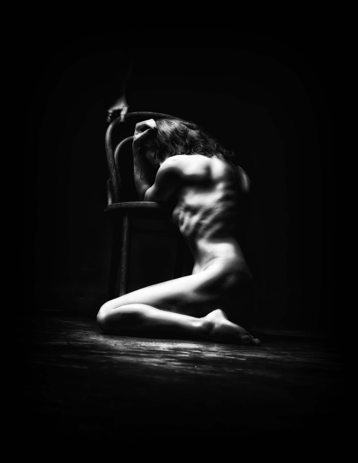 Black and white fine art nude photograph of a figure kneeling on a dark floor beside a chair in dramatic lighting.