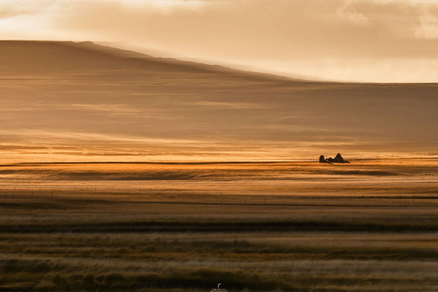 Golden hour sunset over vast open plains with a lone farmhouse silhouetted on the horizon.