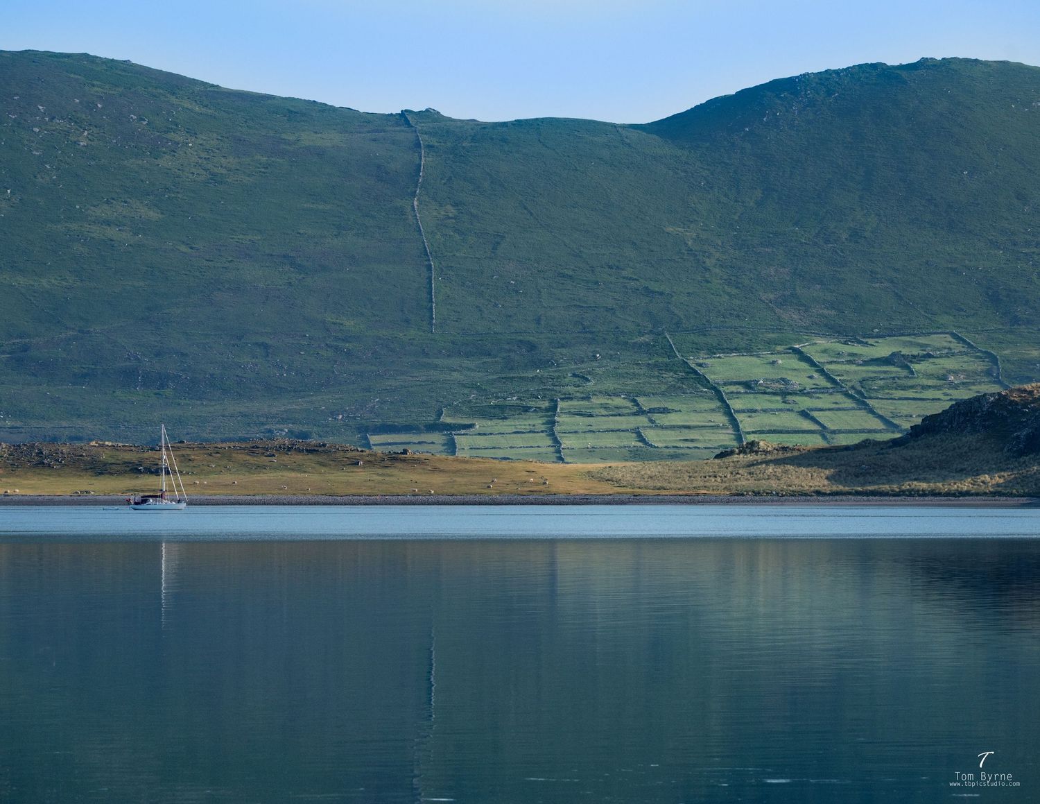 Sailboat on calm Irish loch with green hillside fields and mountain reflections in still blue water.