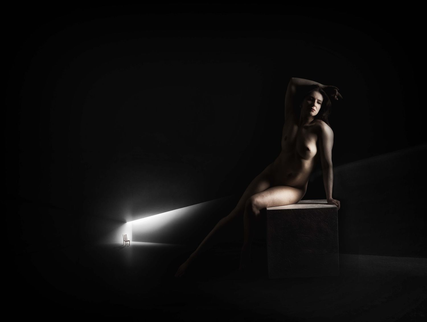 Dramatic fine art nude photography with chiaroscuro lighting, figure seated on stool beside glowing light beam in dark studio.