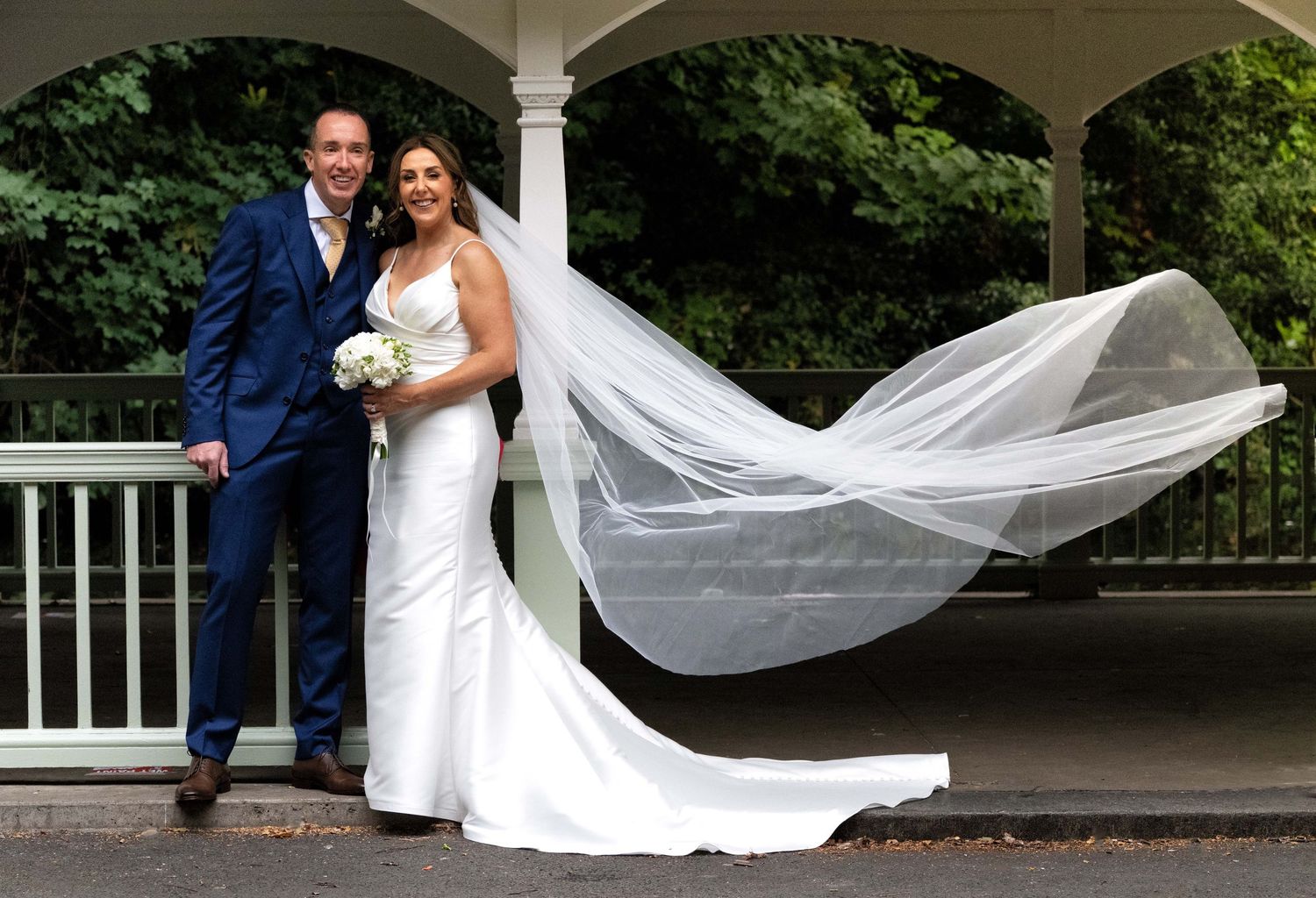 Bride and groom posing outdoors, her veil flowing in the wind, wearing white gown and blue suit near a gazebo.