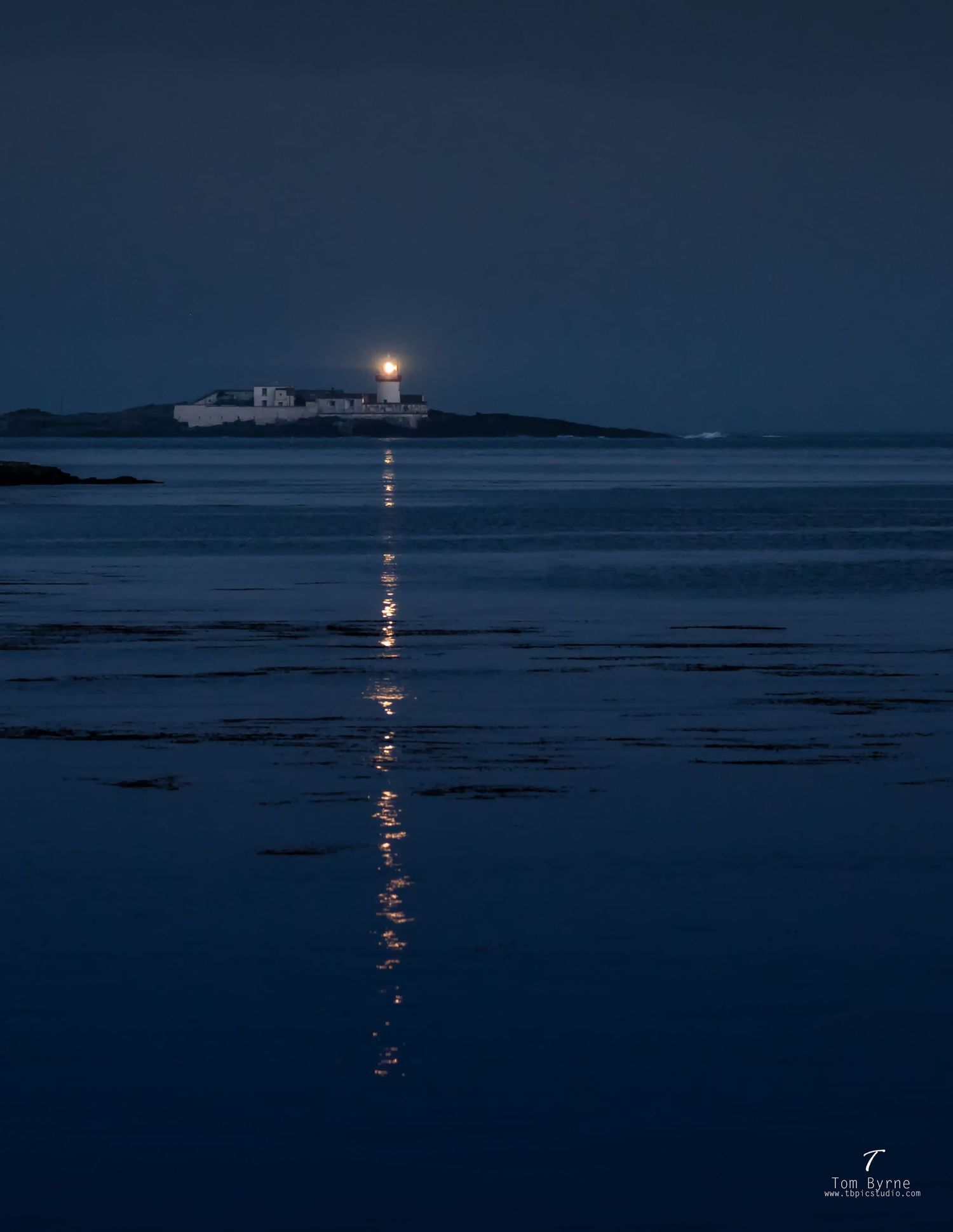 Moonlit lighthouse on a rocky island reflects golden light across calm dark blue ocean waters at night.