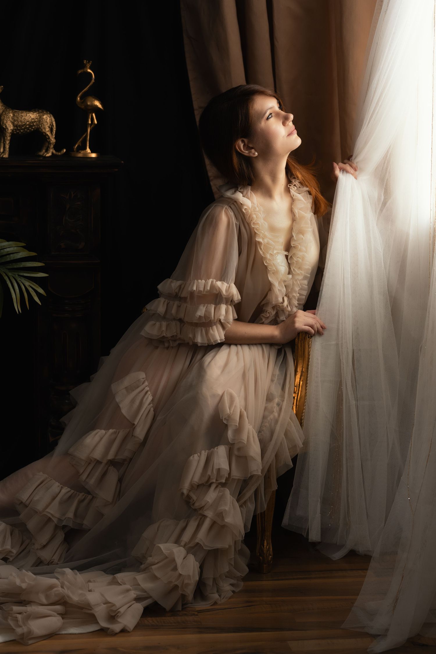 A woman in a vintage ruffled cream gown gazes upward while holding sheer curtains in an elegant, dimly lit room.