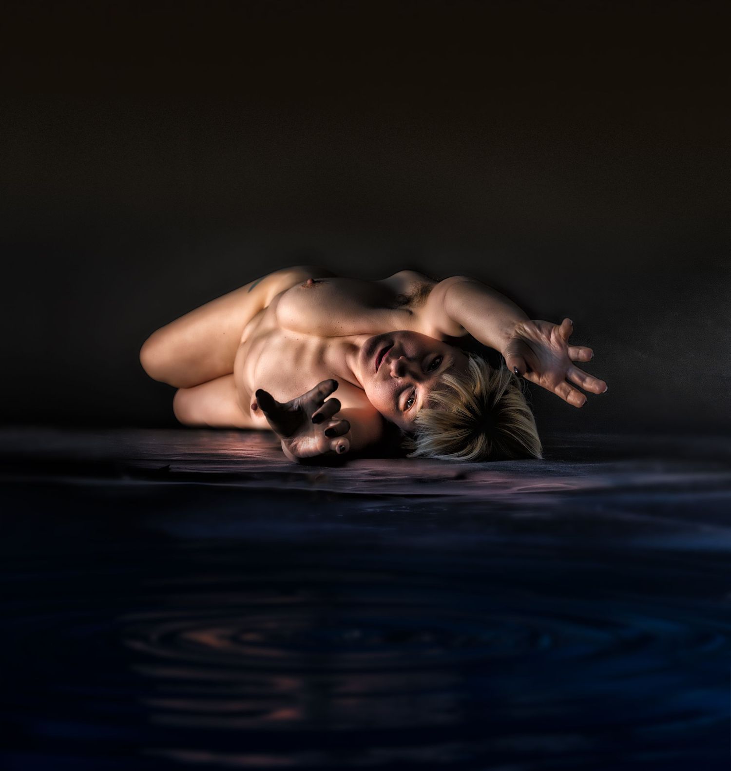 A person lying on a reflective surface in dramatic lighting, reaching outward in an artistic pose.