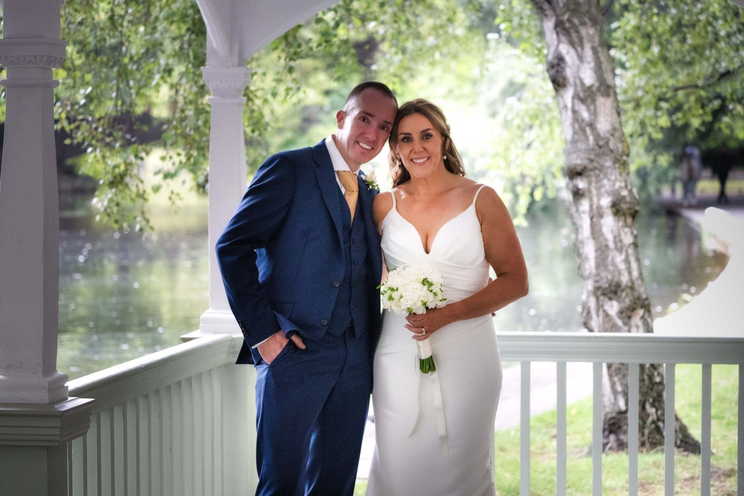 Bride and groom posing on a white gazebo by a pond, groom in navy suit, bride in white gown holding bouquet.