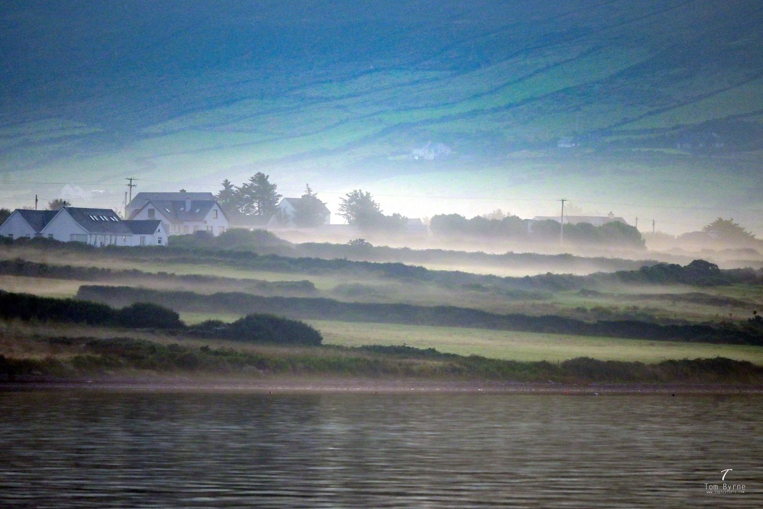 Misty morning landscape with white farmhouse, green fields, and fog rolling over hills near a calm waterway.