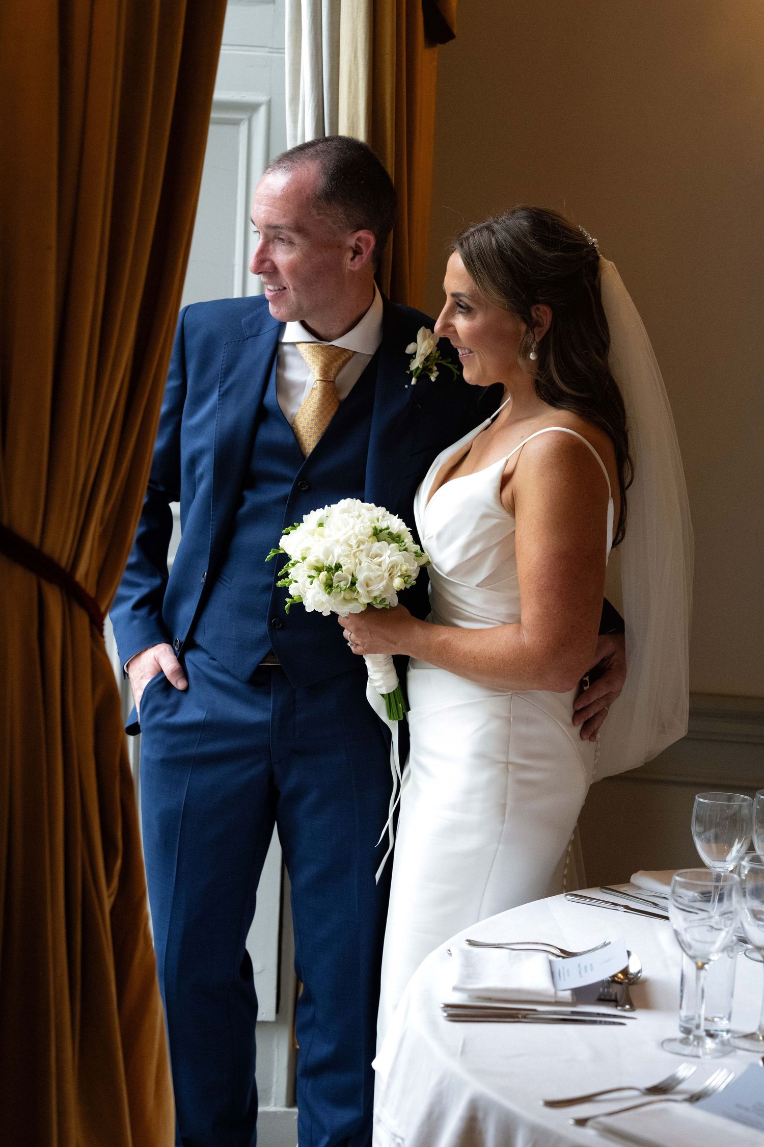 Bride and groom standing together by a window, bride holding white bouquet in elegant white gown and groom in navy suit.