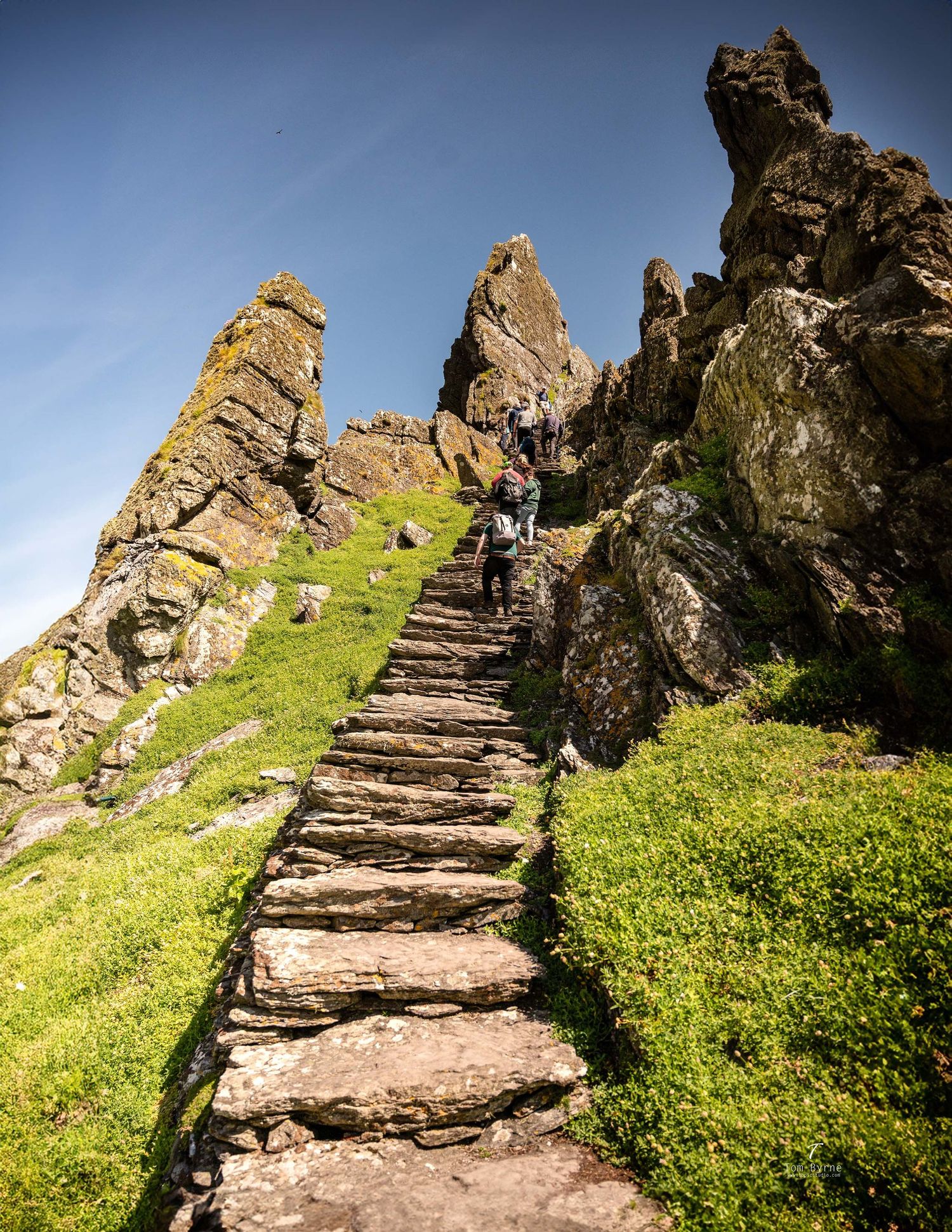 Ancient stone steps wind up a dramatic rocky cliff face under blue skies, with hikers climbing toward the summit.