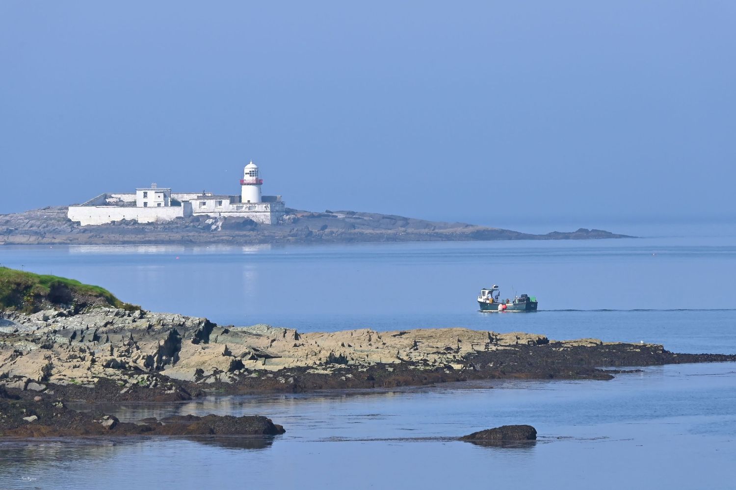 Scenic coastal view with rocky shoreline, calm blue sea, a small boat, and a white lighthouse on a distant island.