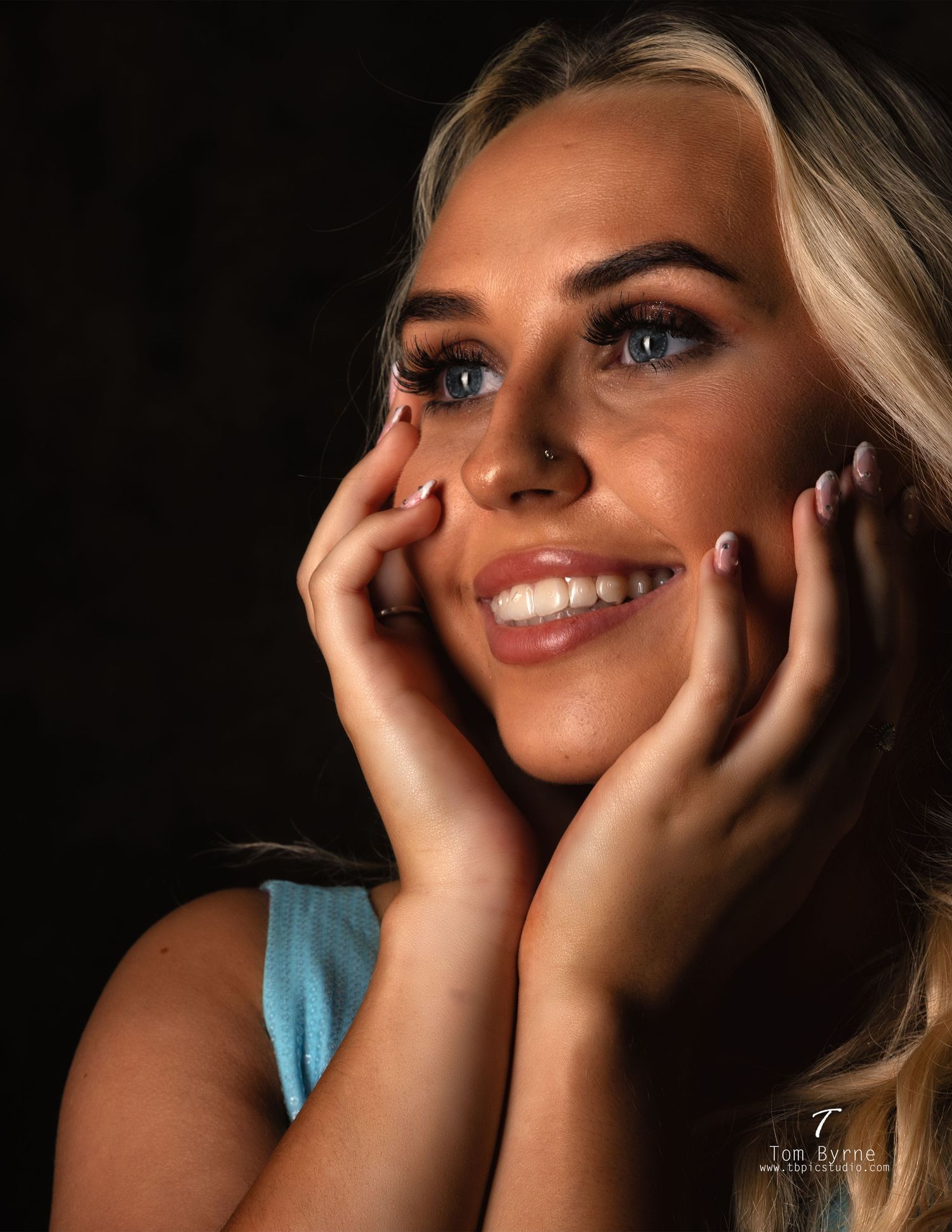 Smiling blonde woman with blue eyes holds her face in her hands against a dark studio background.
