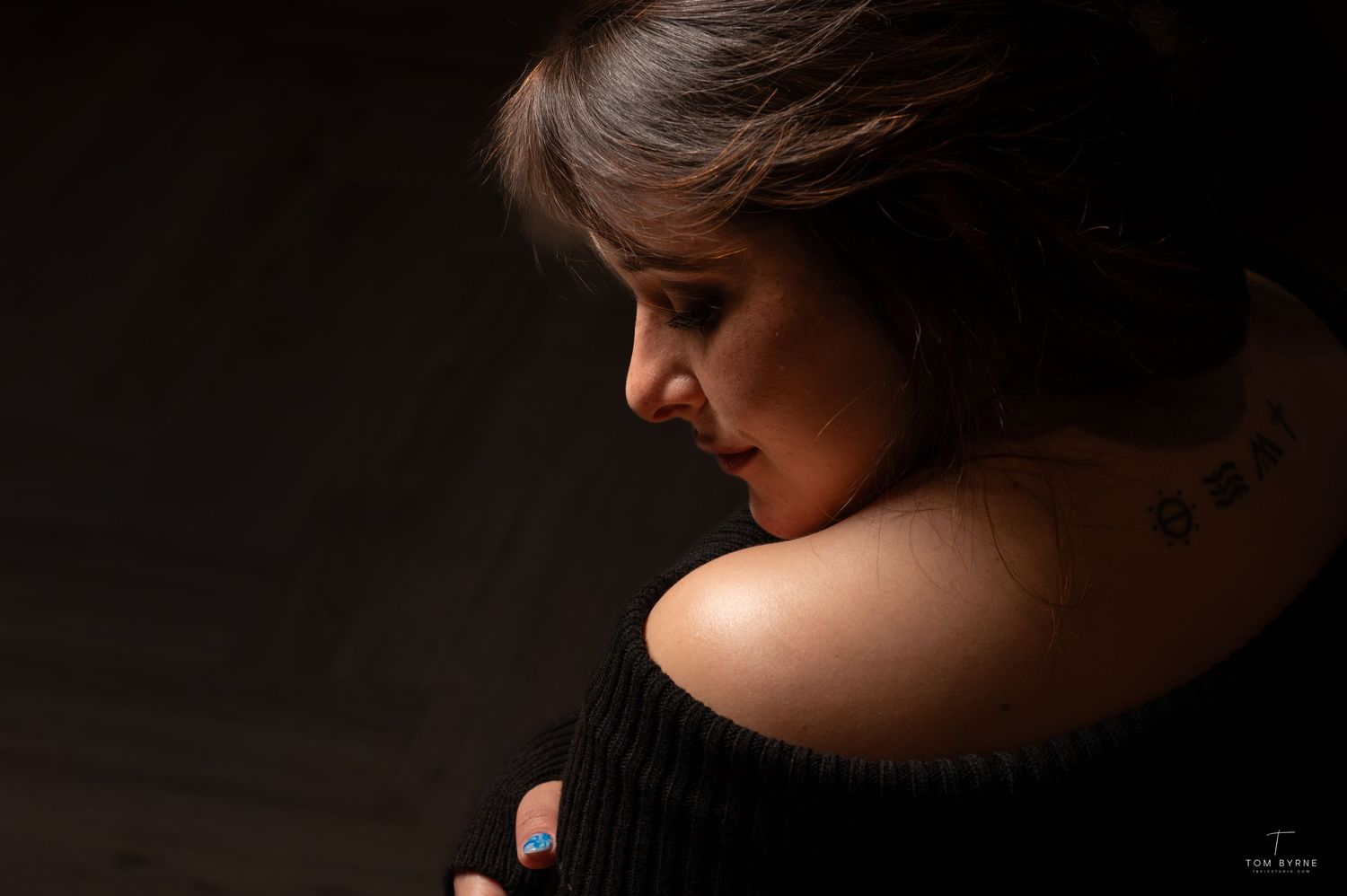 A woman in a black off-shoulder top gazes down, showcasing a neck tattoo in a dramatic studio portrait.