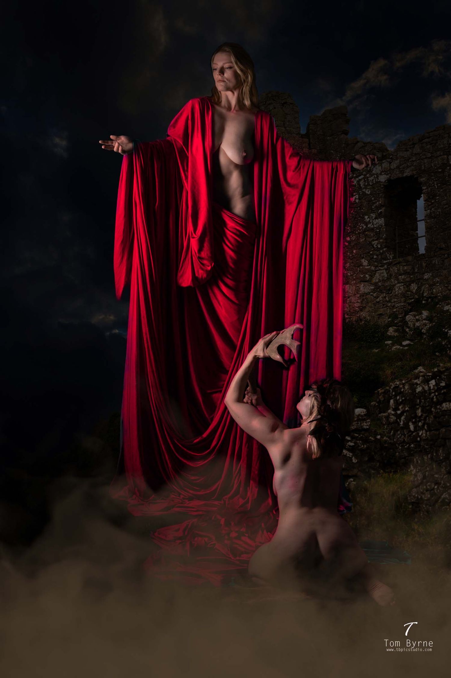 Two figures draped in flowing red fabric pose dramatically against a dark, moody stone ruin backdrop.