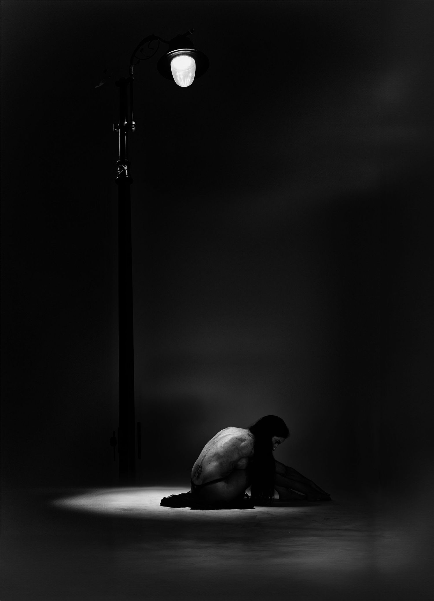 A solitary figure crouches under a glowing street lamp in a dramatic black and white fine art photograph.