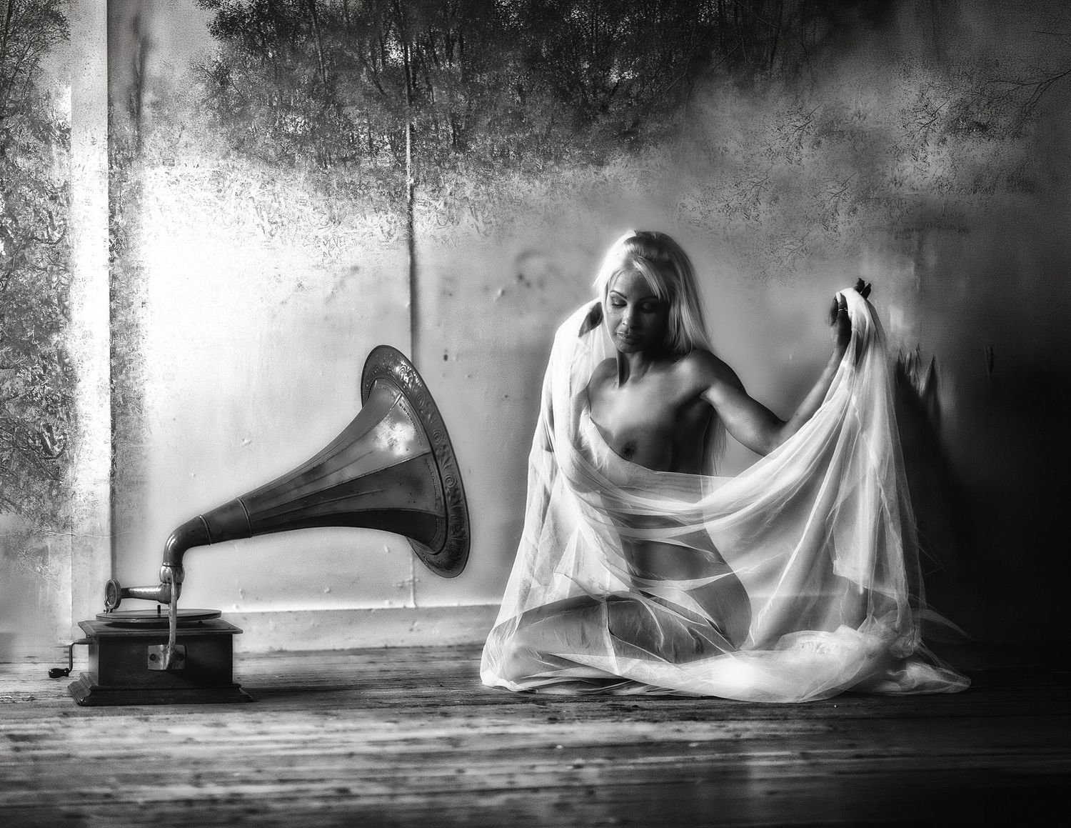 Black and white photo of a woman draped in sheer fabric sitting beside a vintage gramophone on a wooden floor.