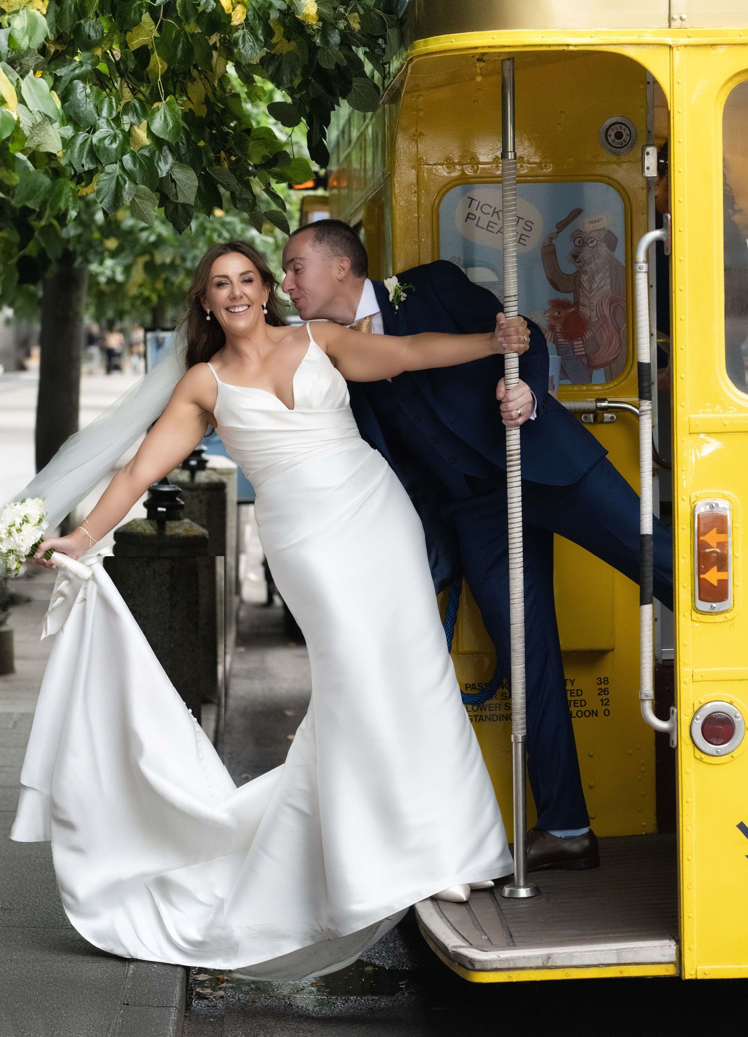 Bride in white gown and groom in navy suit pose on yellow bus steps on wedding day.