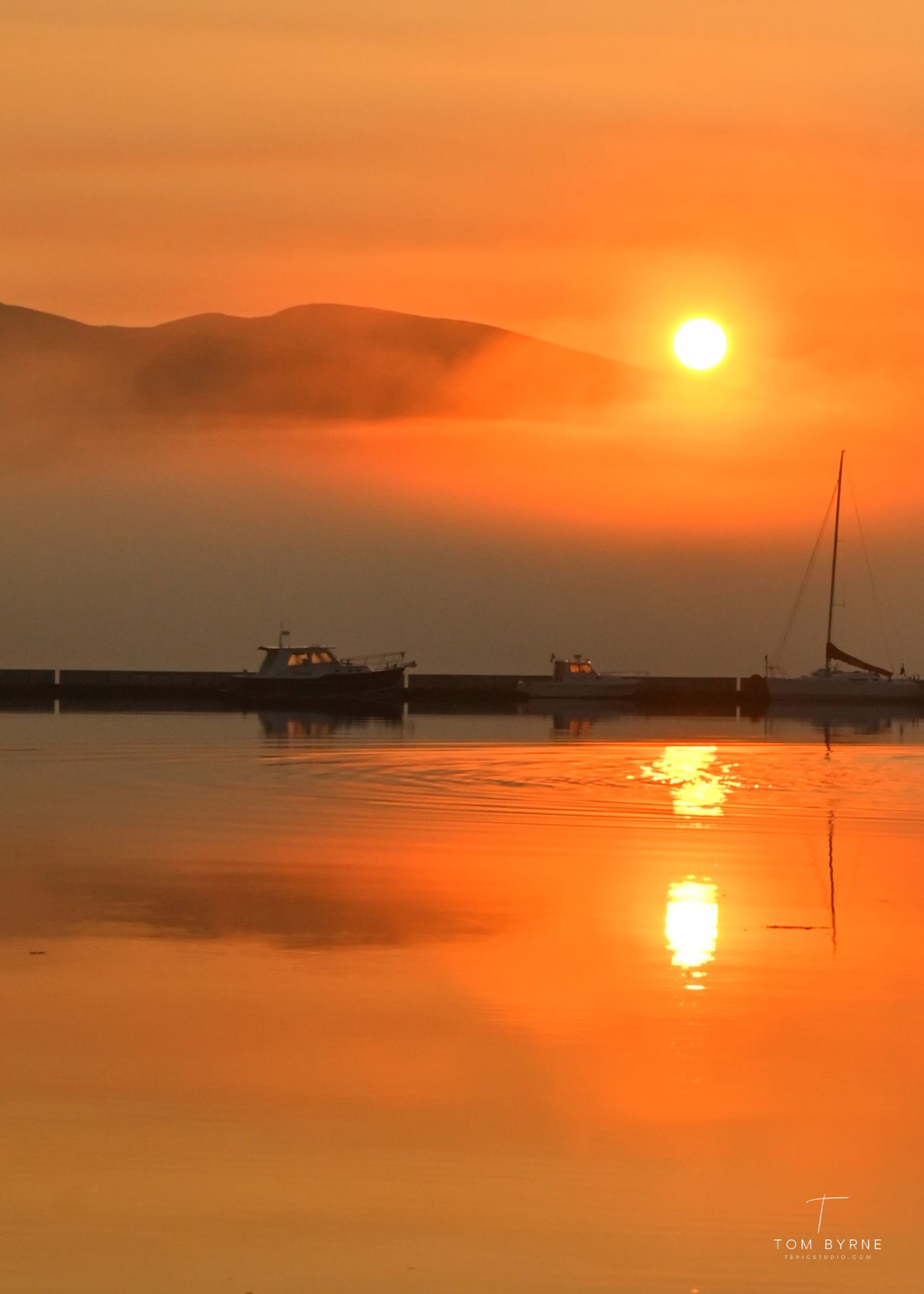 Golden sunset over a misty harbor with boats and sailboat reflections on calm orange water.