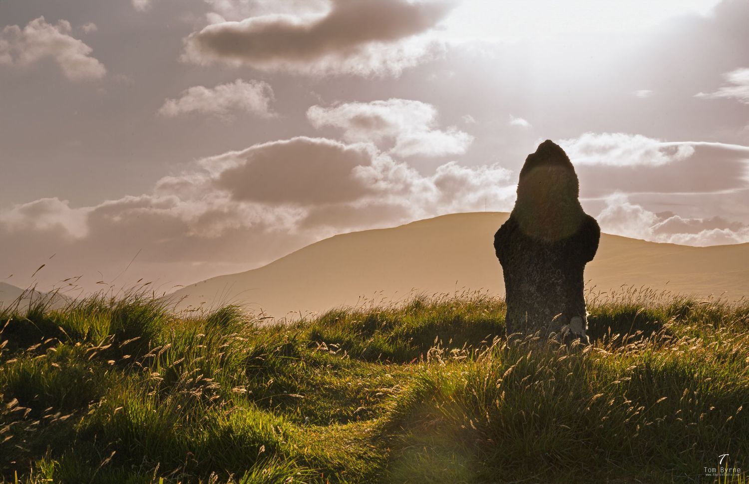 Ancient standing stone monument in lush green grass field with dramatic cloudy sky and rolling hills at sunset.