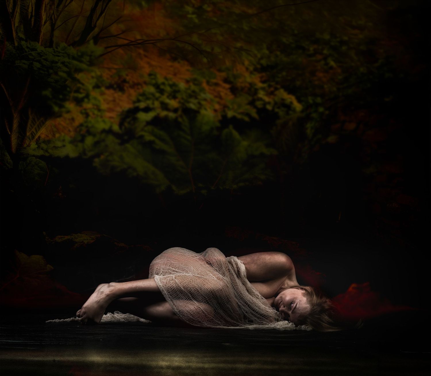 A woman draped in sheer fabric lies curled on a dark surface beneath autumn foliage in moody, dramatic lighting.