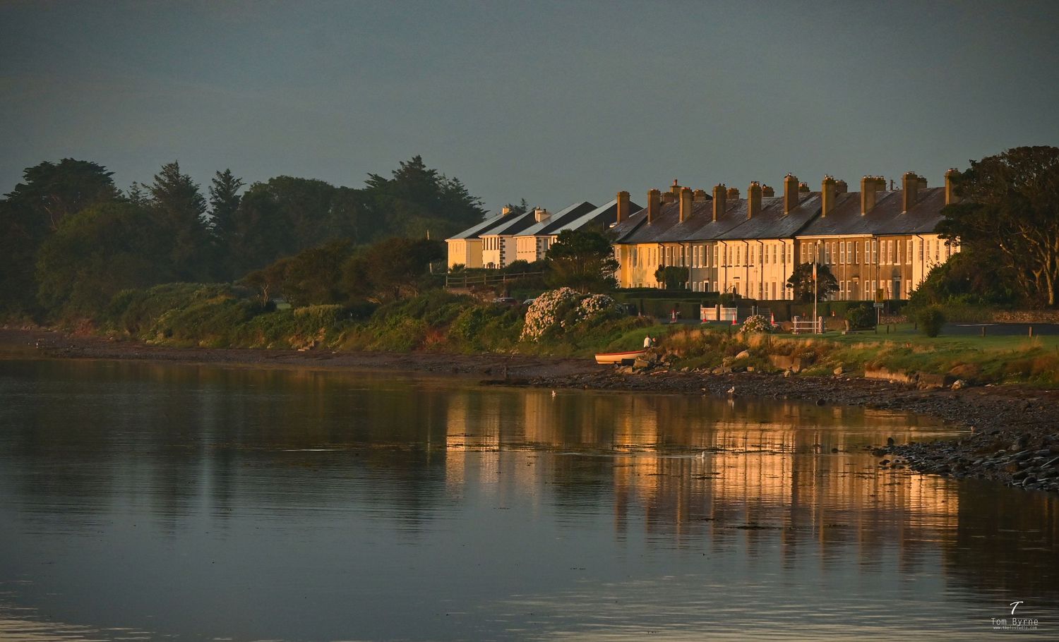 Golden hour light illuminates historic waterfront cottages reflecting in calm estuary waters, surrounded by lush green trees.