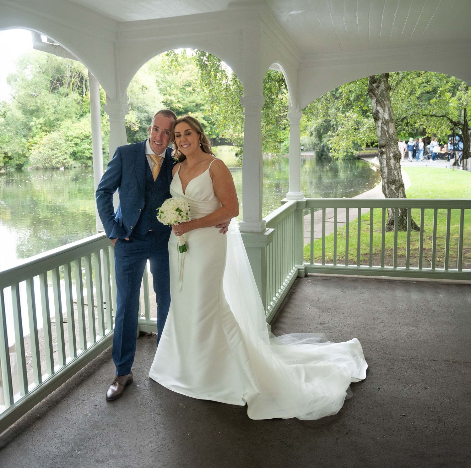 Bride and groom posing in a white gazebo overlooking a serene lake, surrounded by lush green trees.