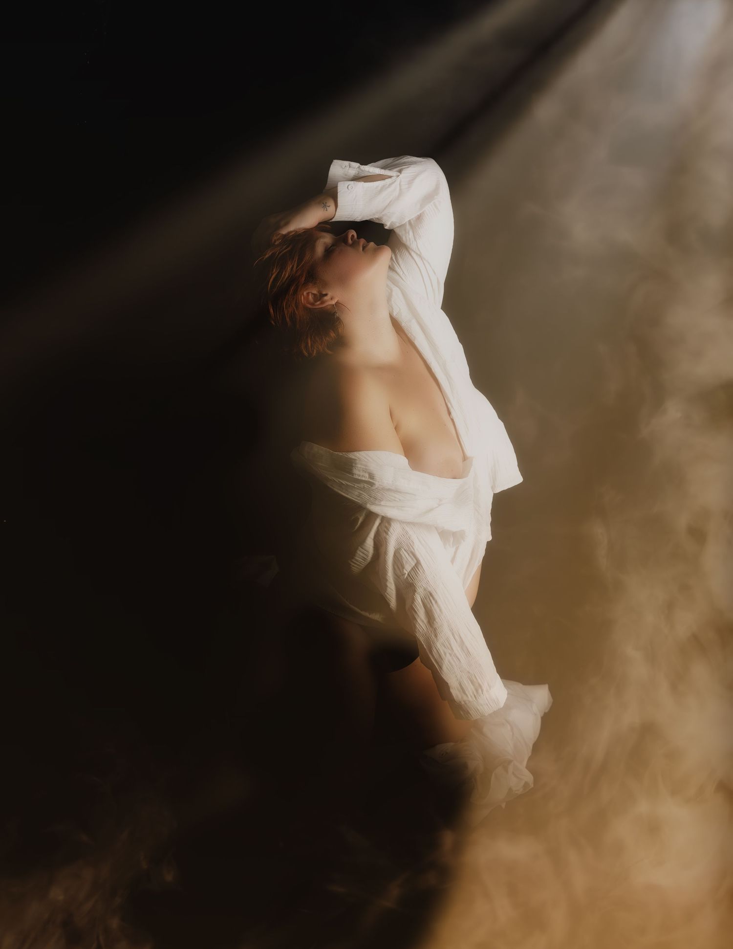 A person in a flowing white off-shoulder garment poses dramatically in a beam of light amid golden smoke.