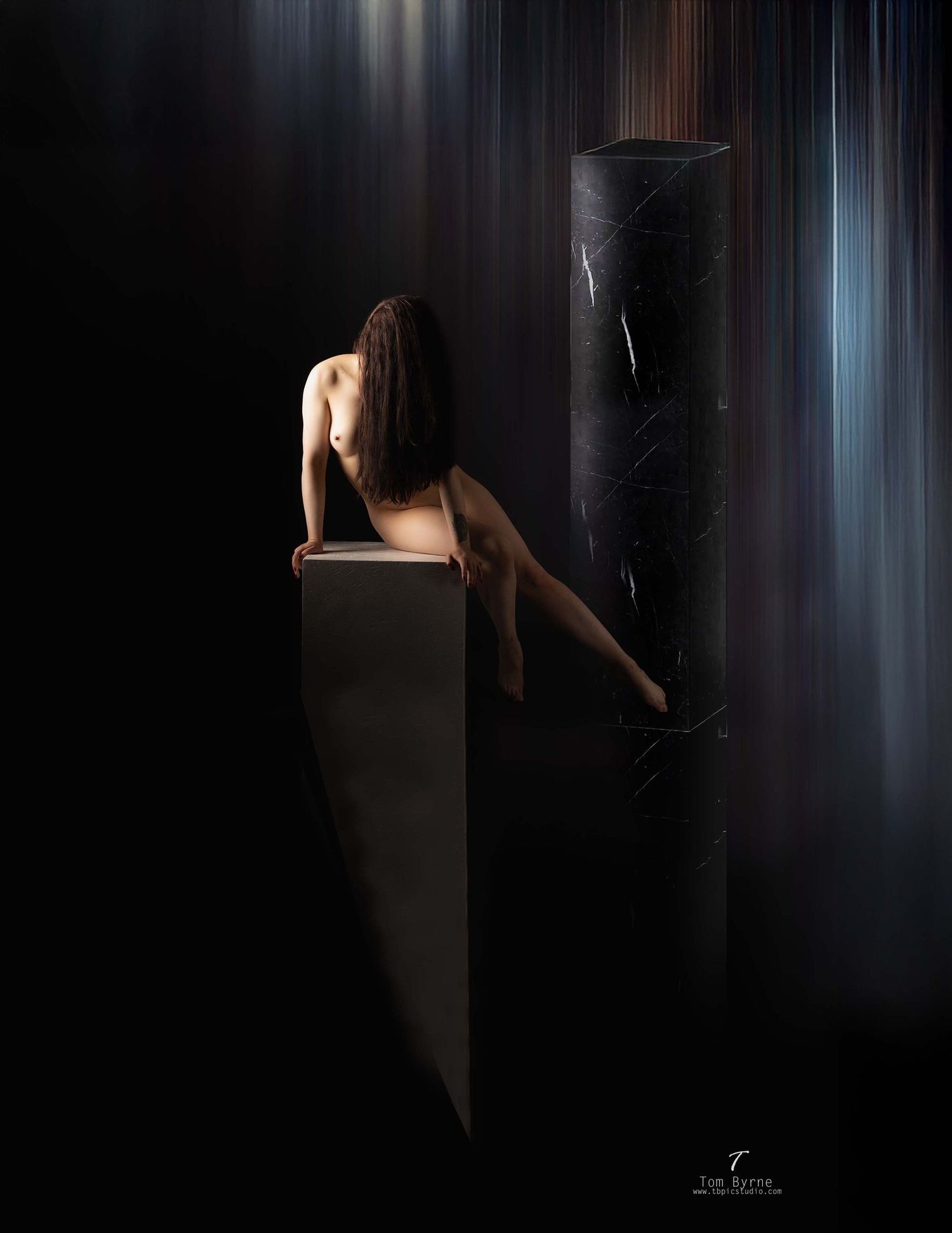 Artistic nude figure perched on a dark pedestal beside a tall black column with dramatic light streaks in background.