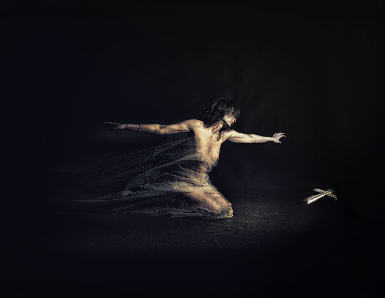 A dancer kneels dramatically in darkness, arms outstretched toward a small glowing object on the ground.