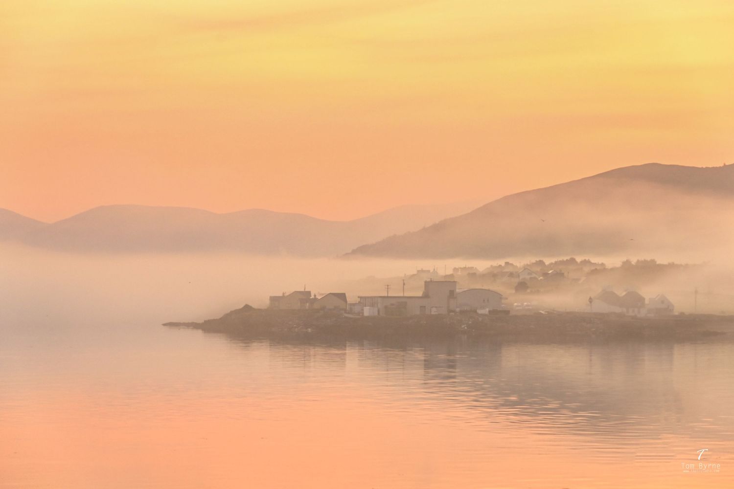 Misty golden sunrise over a tranquil lake with a small village on a peninsula, surrounded by rolling hills in soft orange hues.