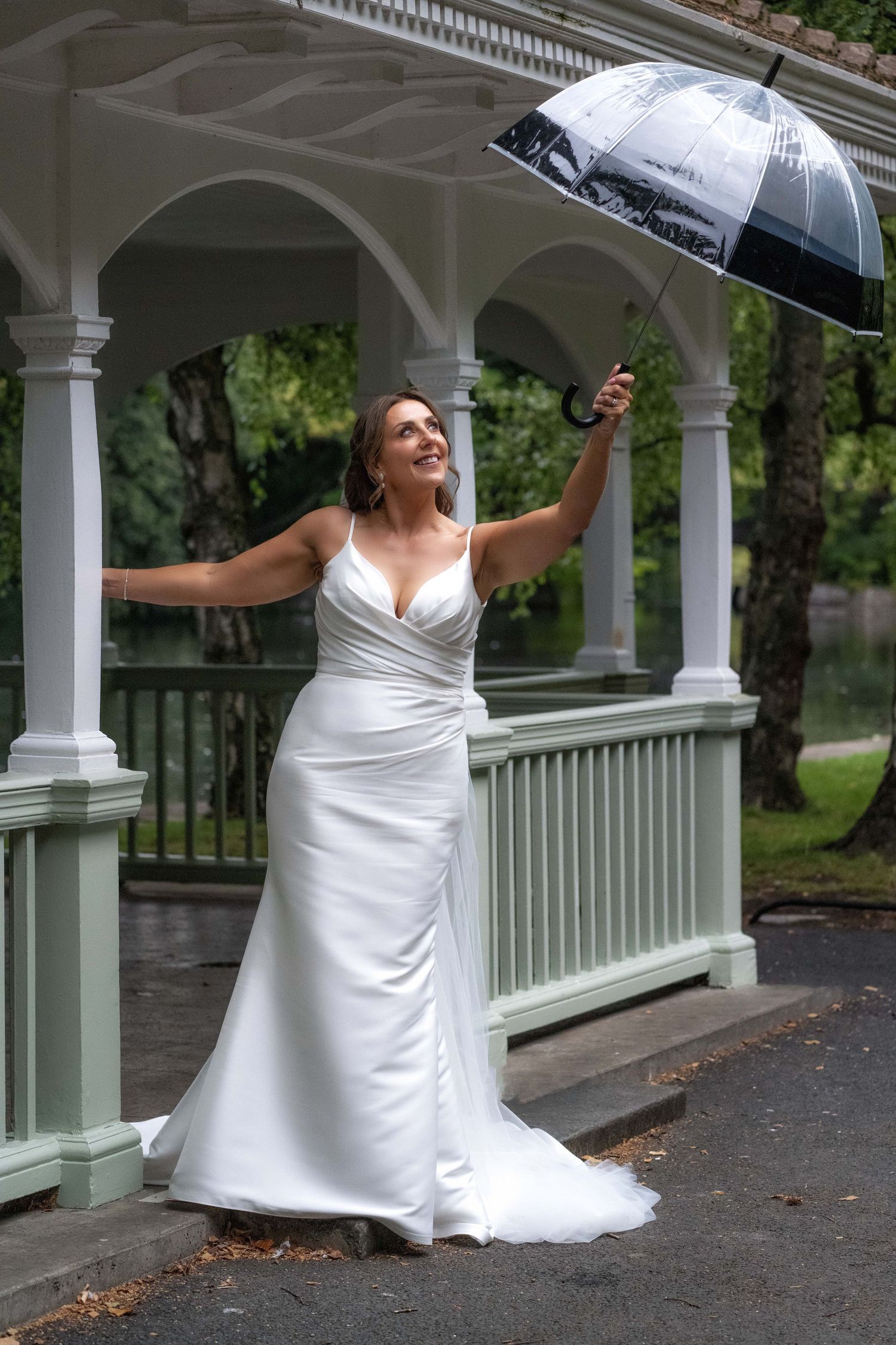 Bride in white wedding gown holding clear umbrella, smiling joyfully beside a white gazebo on a rainy day.