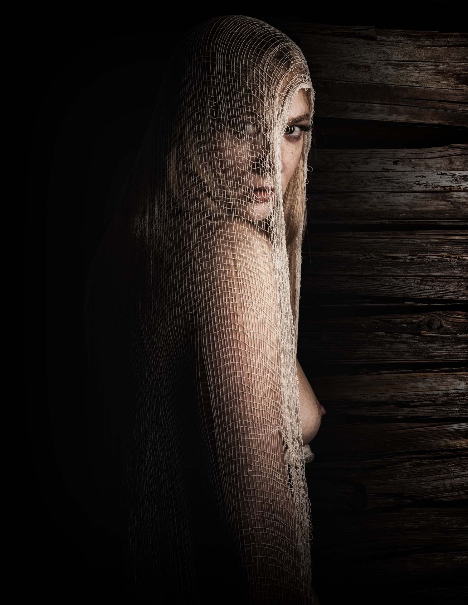 A mysterious woman draped in sheer mesh veil against dark wooden background in dramatic lighting.