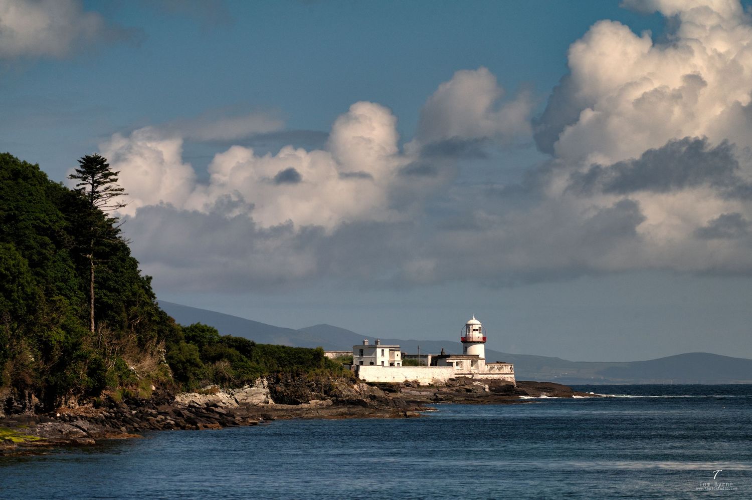 White lighthouse on rocky Irish coastline with lush green trees and dramatic cloudy sky over calm blue waters.