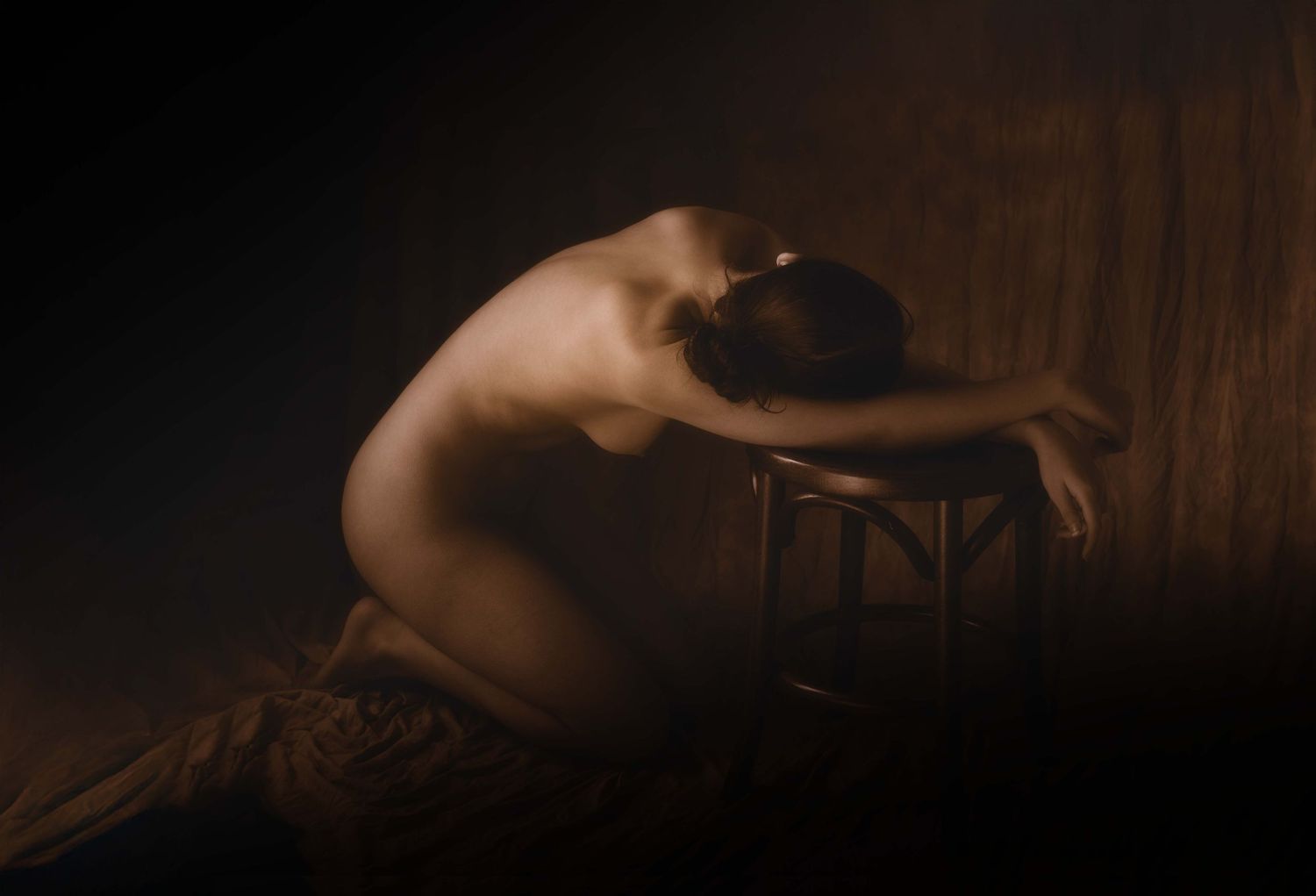 A nude figure kneels in shadow, head resting on arms draped over a wooden stool, in dramatic chiaroscuro lighting.